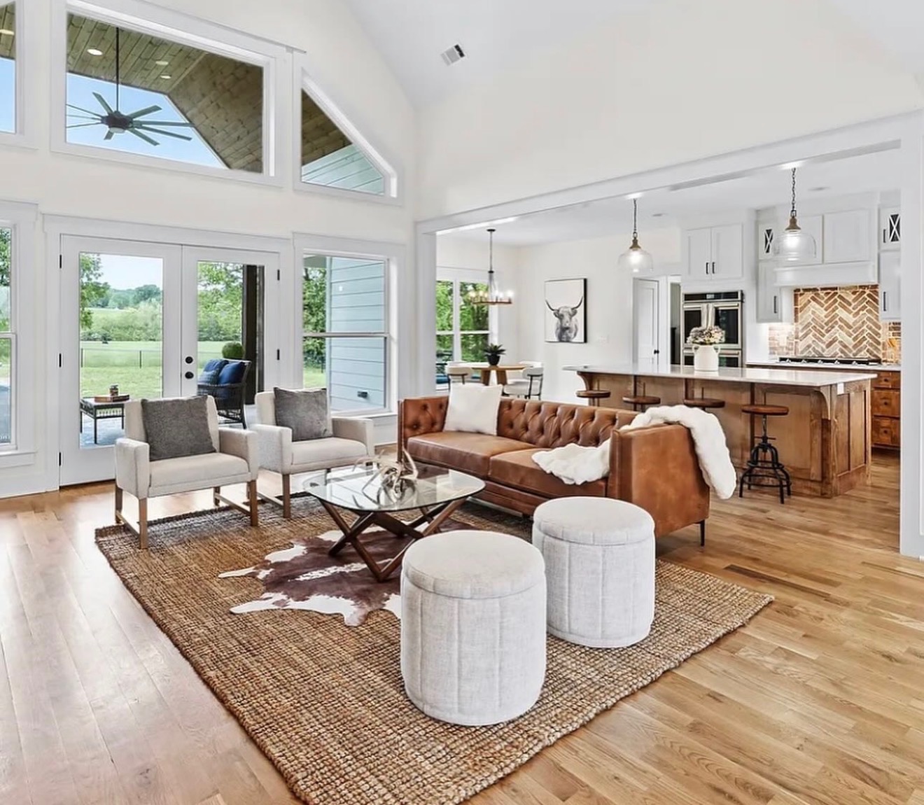 Living room with large windows, brown sofa, white chairs, and a kitchen in the background, bright and spacious interior.