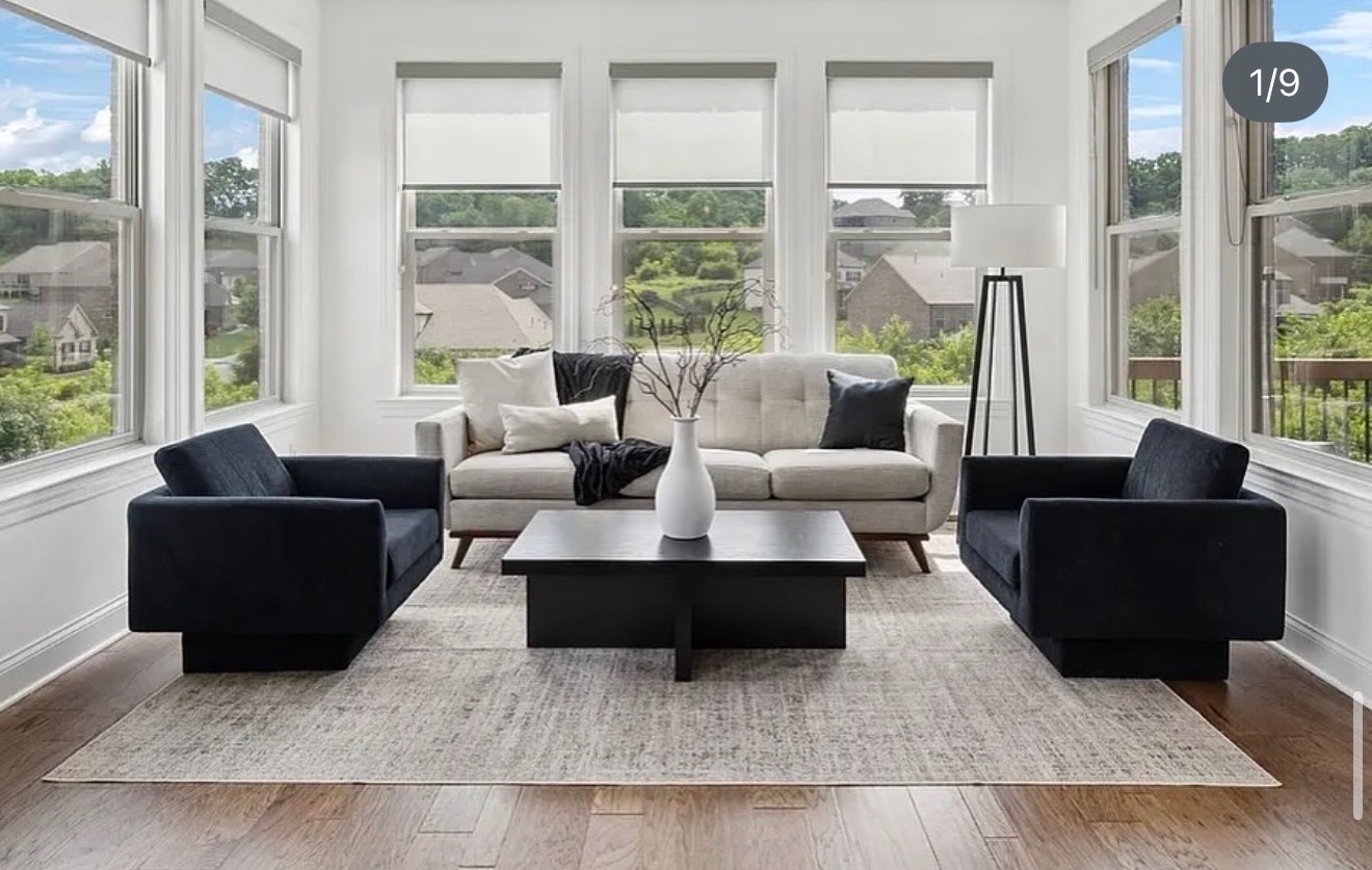Living room with large windows, white sofa, two black armchairs, a black coffee table, and a floor lamp, with a view of greenery outside.