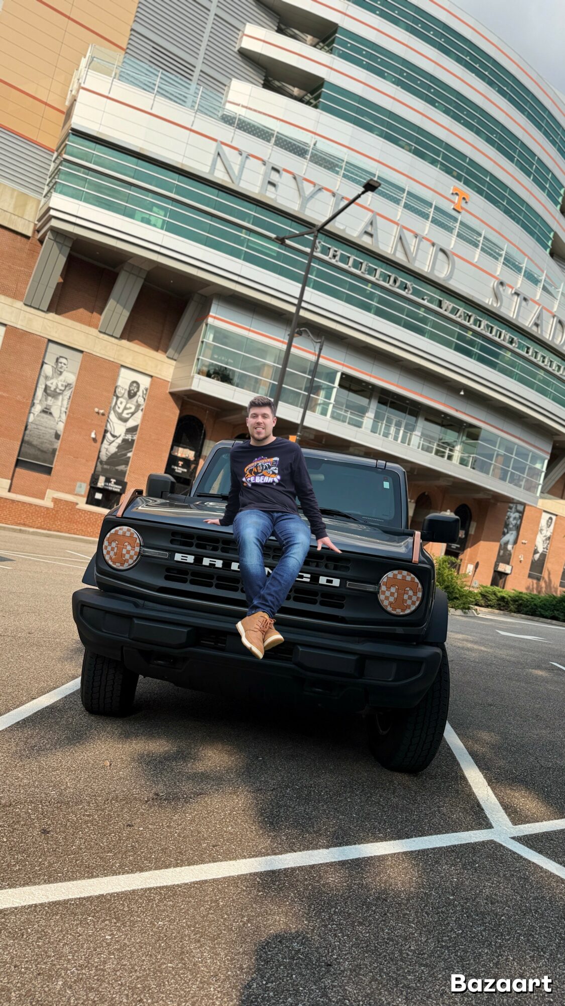 Person sitting on the hood of a black vehicle in front of a modern building with glass windows and a sign reading 'NEYLAND'.