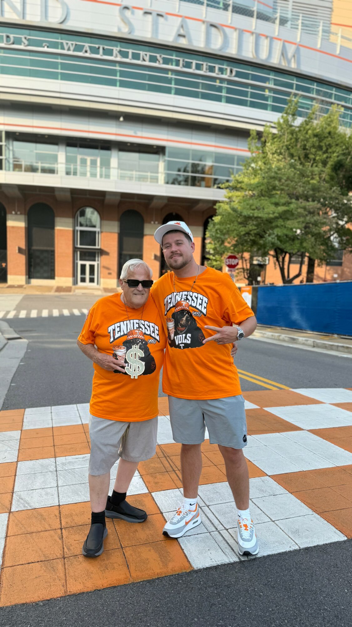 Two people wearing orange T-shirts and shorts standing on a checkered sidewalk in front of a stadium, smiling.