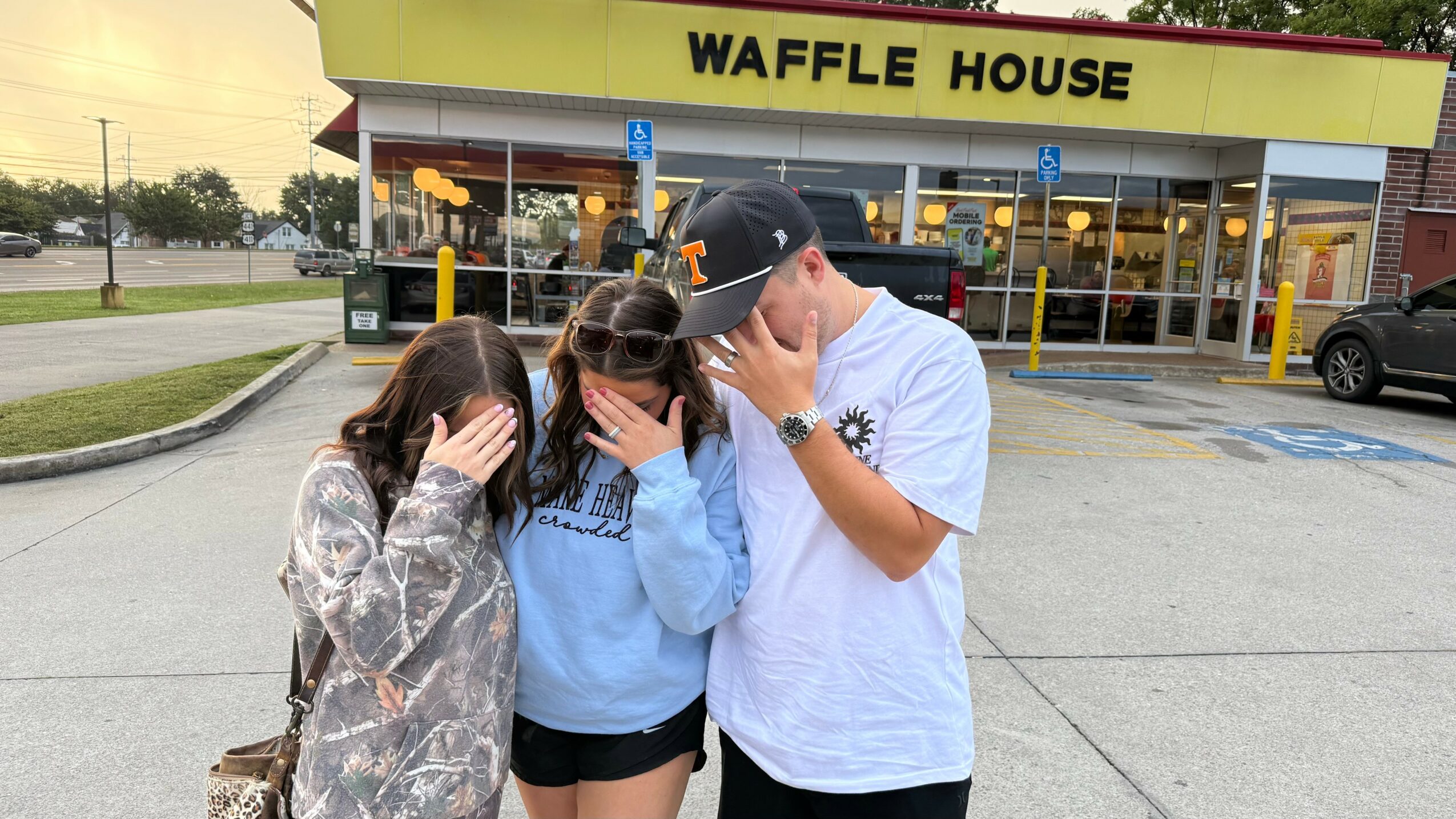 Four people standing outside a Waffle House, covering their faces with their hands, in a parking lot.