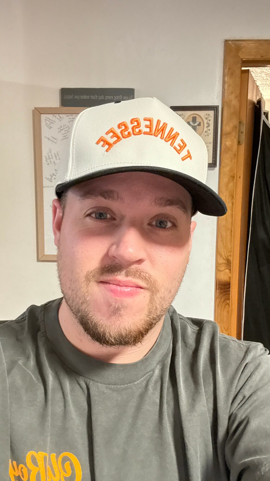 Young man with a beard wearing a white cap with orange text and a gray shirt, smiling indoors.