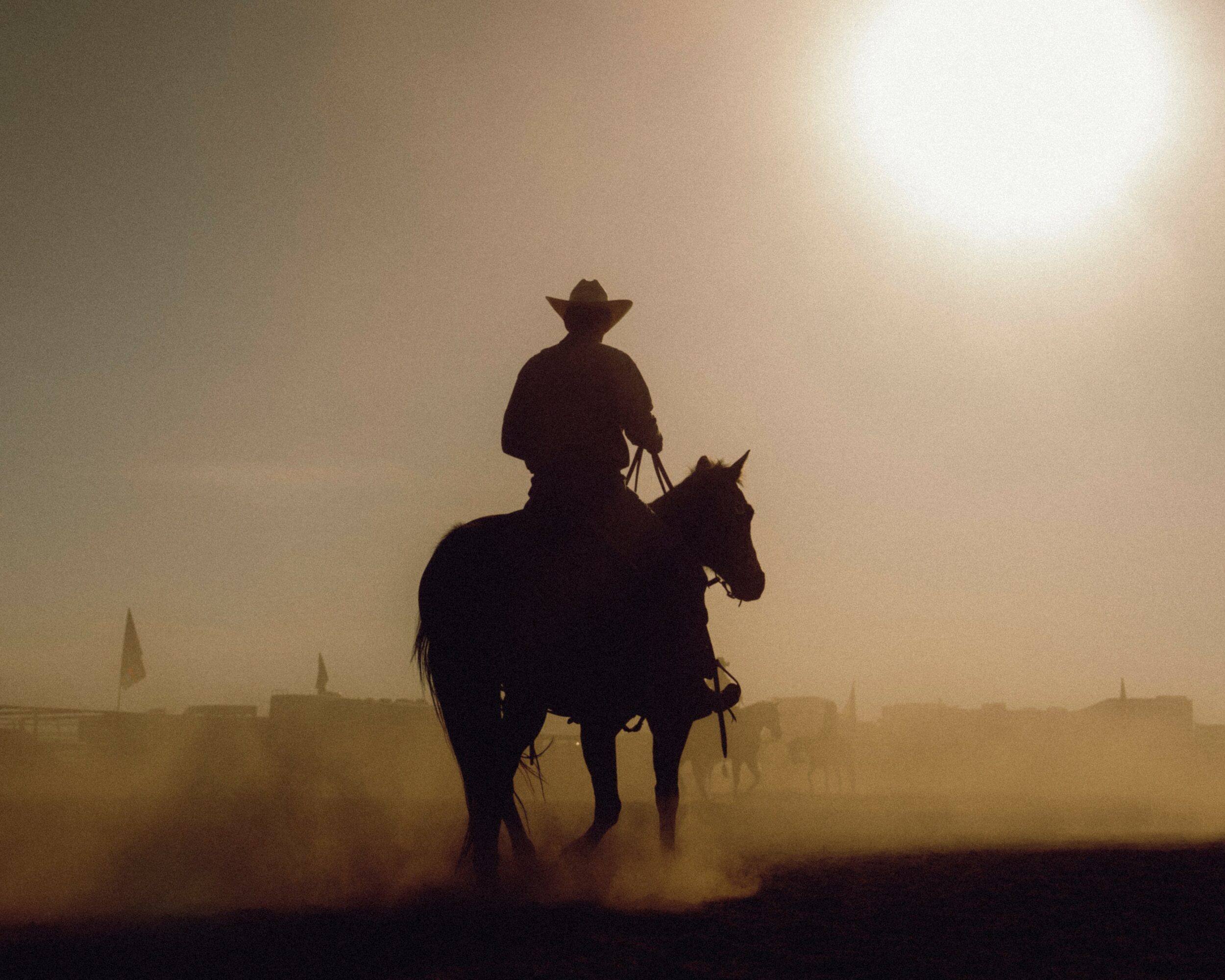 Person riding a horse with a hat, silhouetted against a bright sun, dust in the air, and a hazy background.