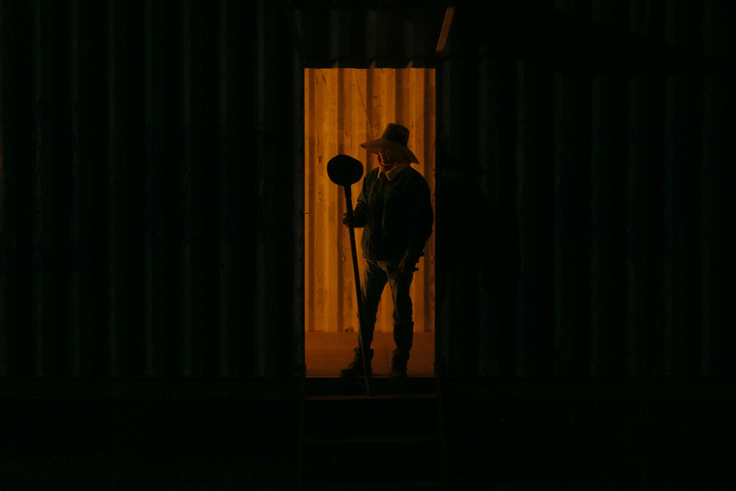Person standing in doorway with warm light behind, holding a hat, silhouette visible against the illuminated background.