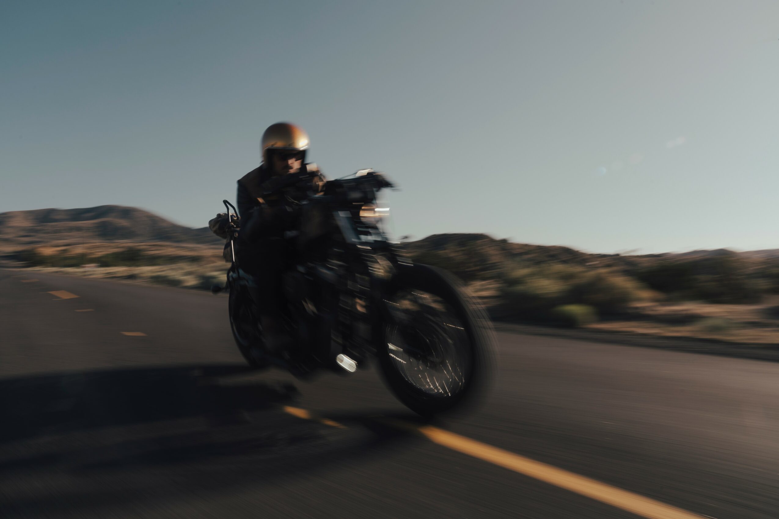 Motorcycle rider wearing helmet and dark clothing riding on a road with desert landscape, motion blurred.