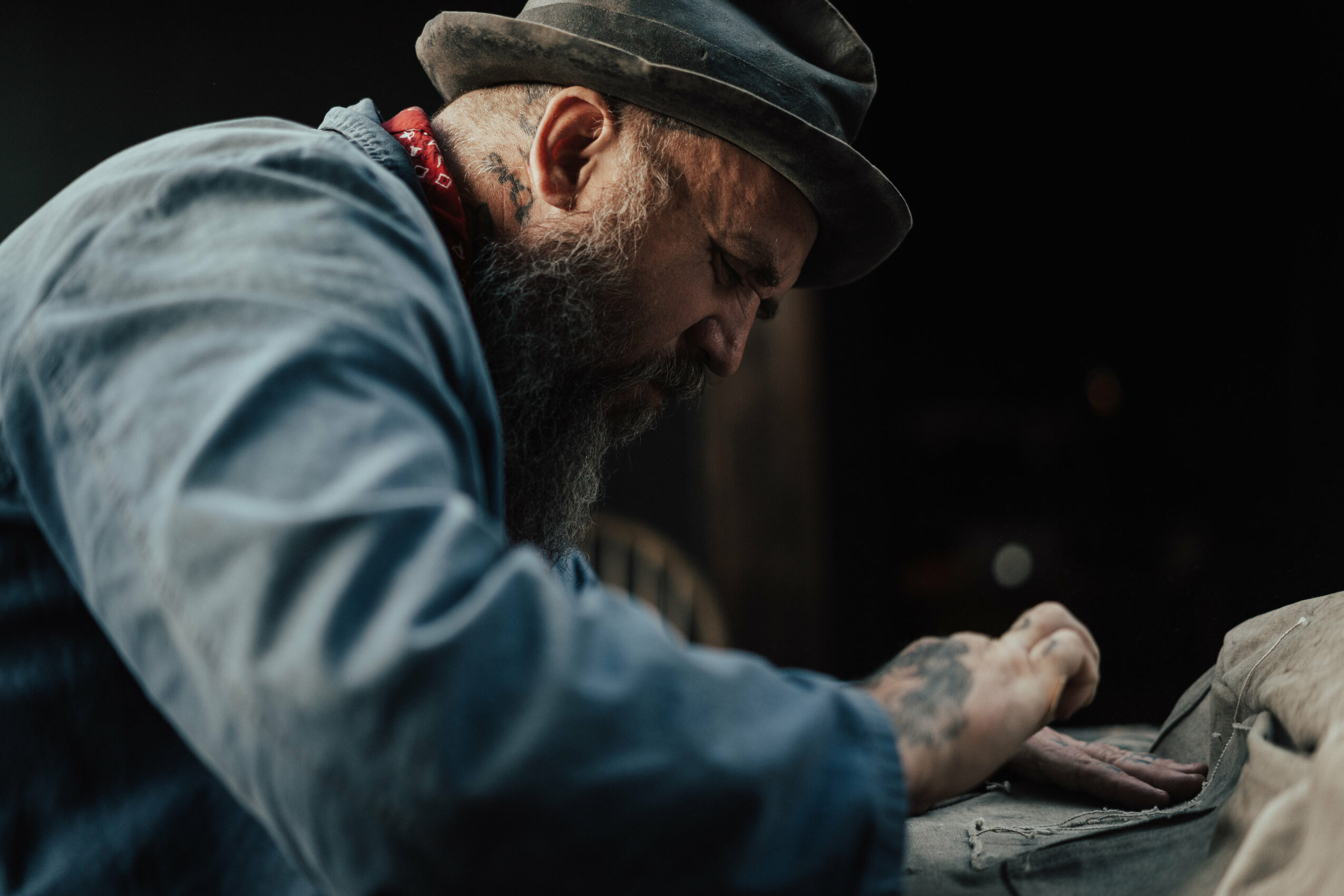 Man with a beard wearing a hat and blue shirt, focused on a task, in a dimly lit environment.