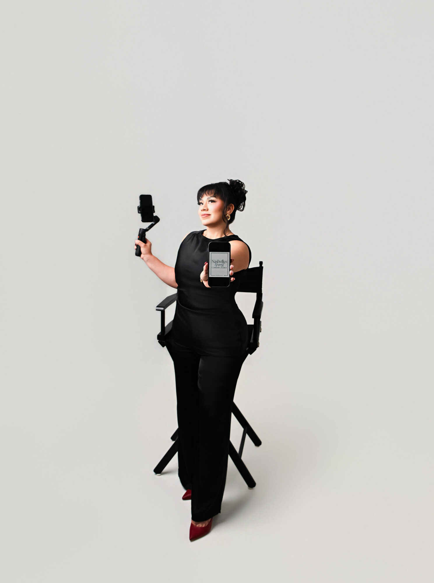 Woman in black dress sitting on a chair, holding a camera and a smartphone, against a plain background.