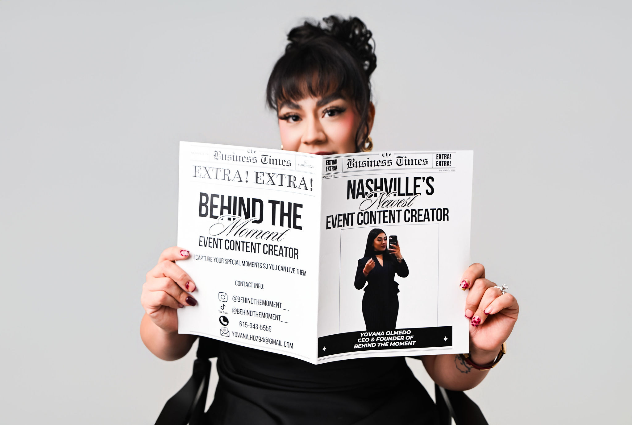 Woman with dark hair and bangs reading a newspaper, holding it open in front of her face, against a plain background.