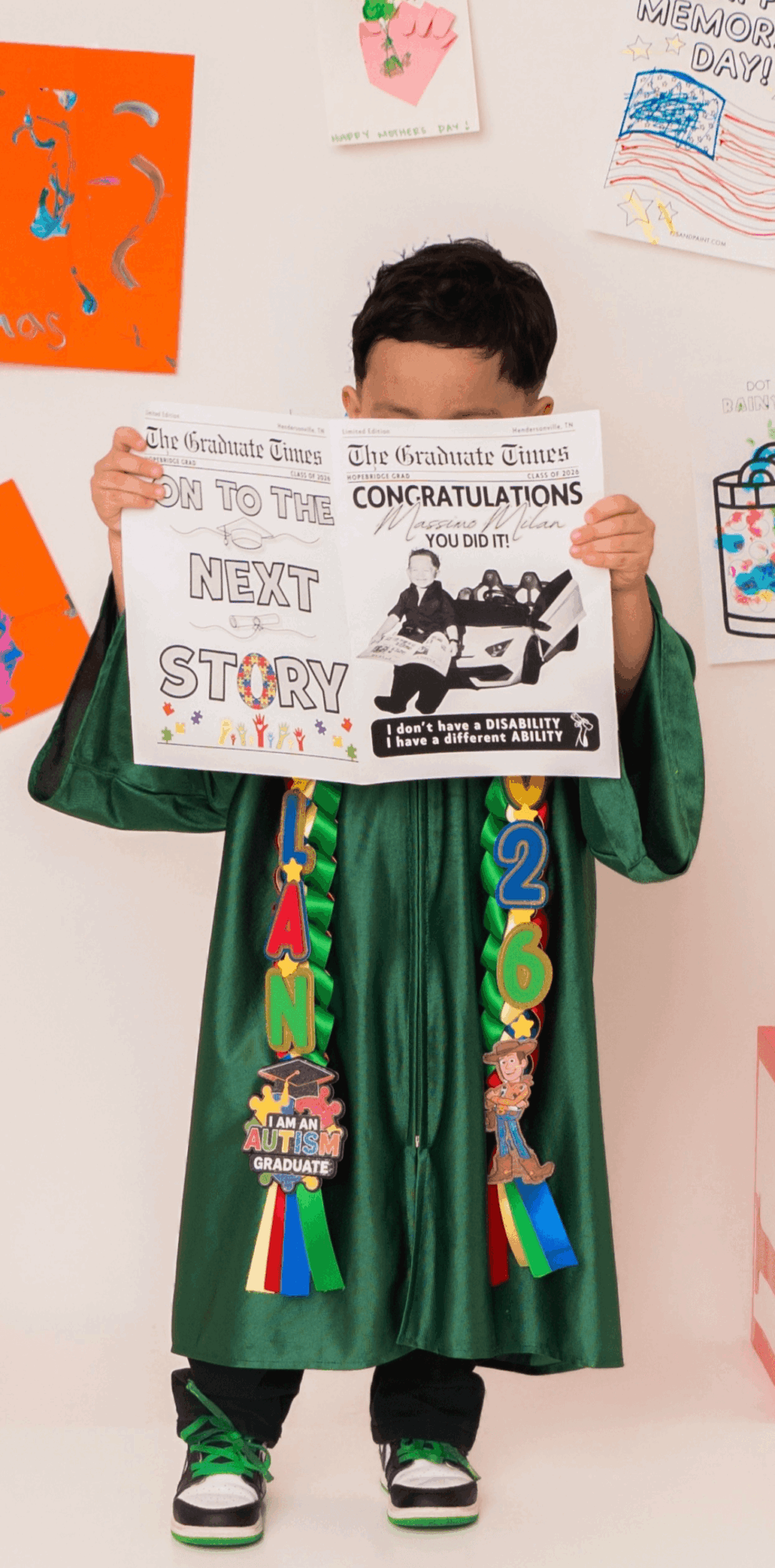 Child in green graduation gown holding a congratulatory sign, decorated with ribbons and medals, standing in front of colorful wall displays.