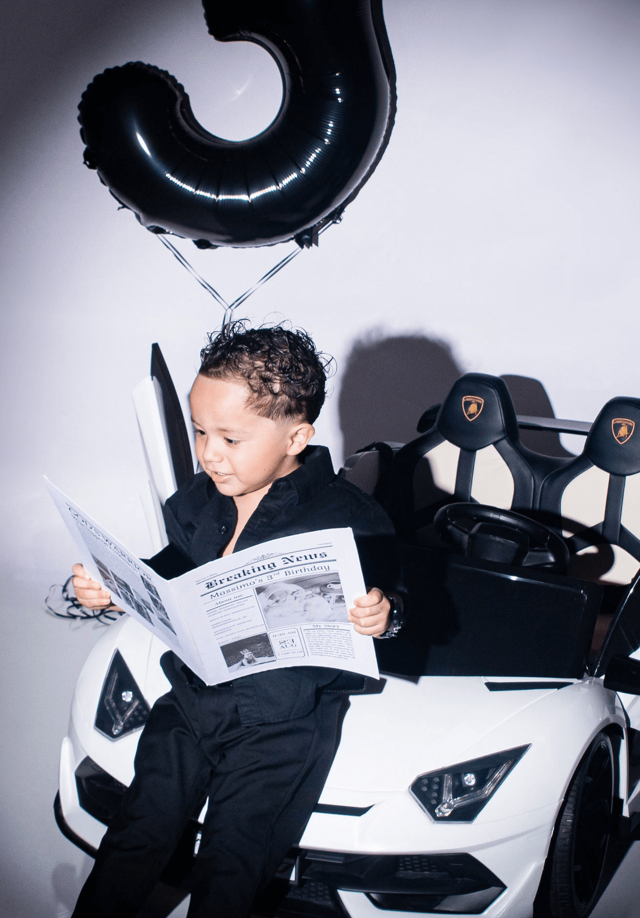 Child sitting in a toy car, reading a newspaper, with a black balloon shaped like the number 3 floating above.