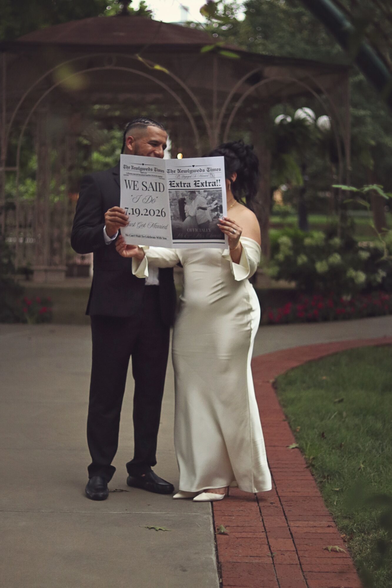 A couple in formal attire holding a newspaper in a park with trees and a gazebo in the background.
