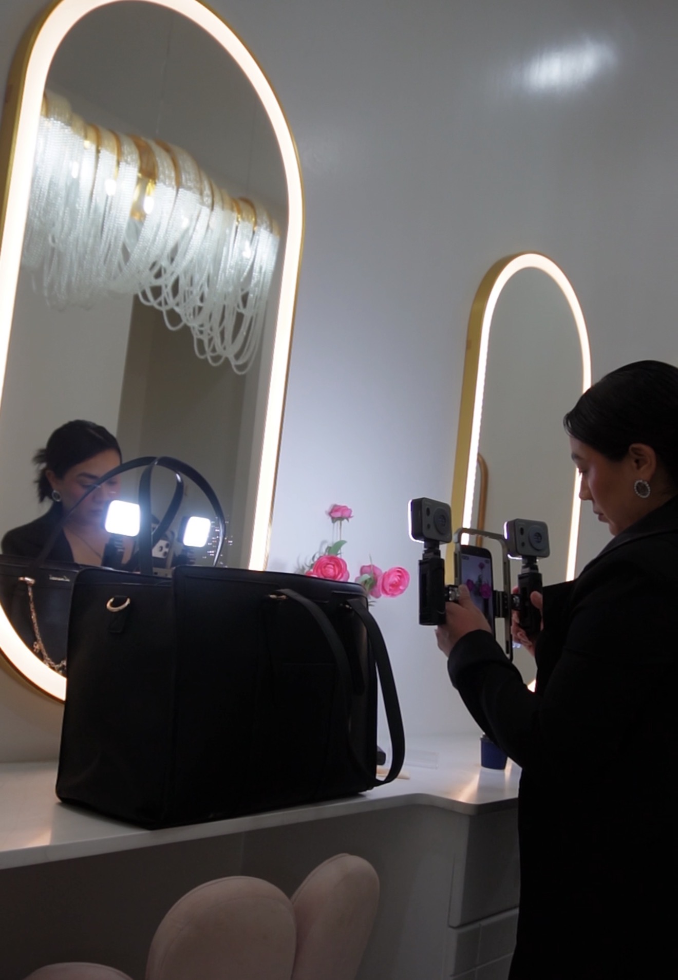 Person taking a photo in front of a mirror with illuminated frames, with a black bag on the counter, pink flowers reflected.