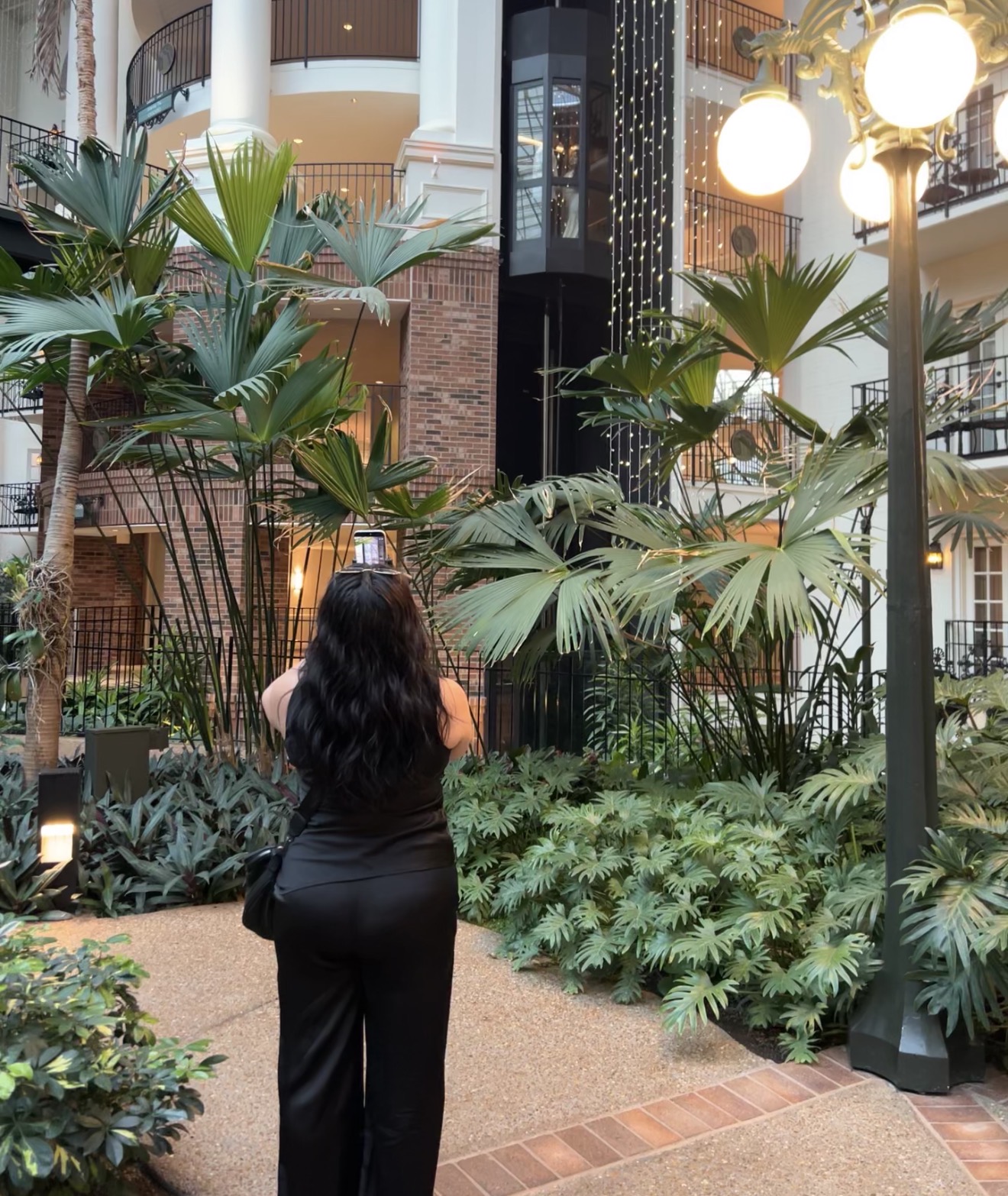 Person with long dark hair standing in an indoor garden area with large green plants and a tall lamp post.