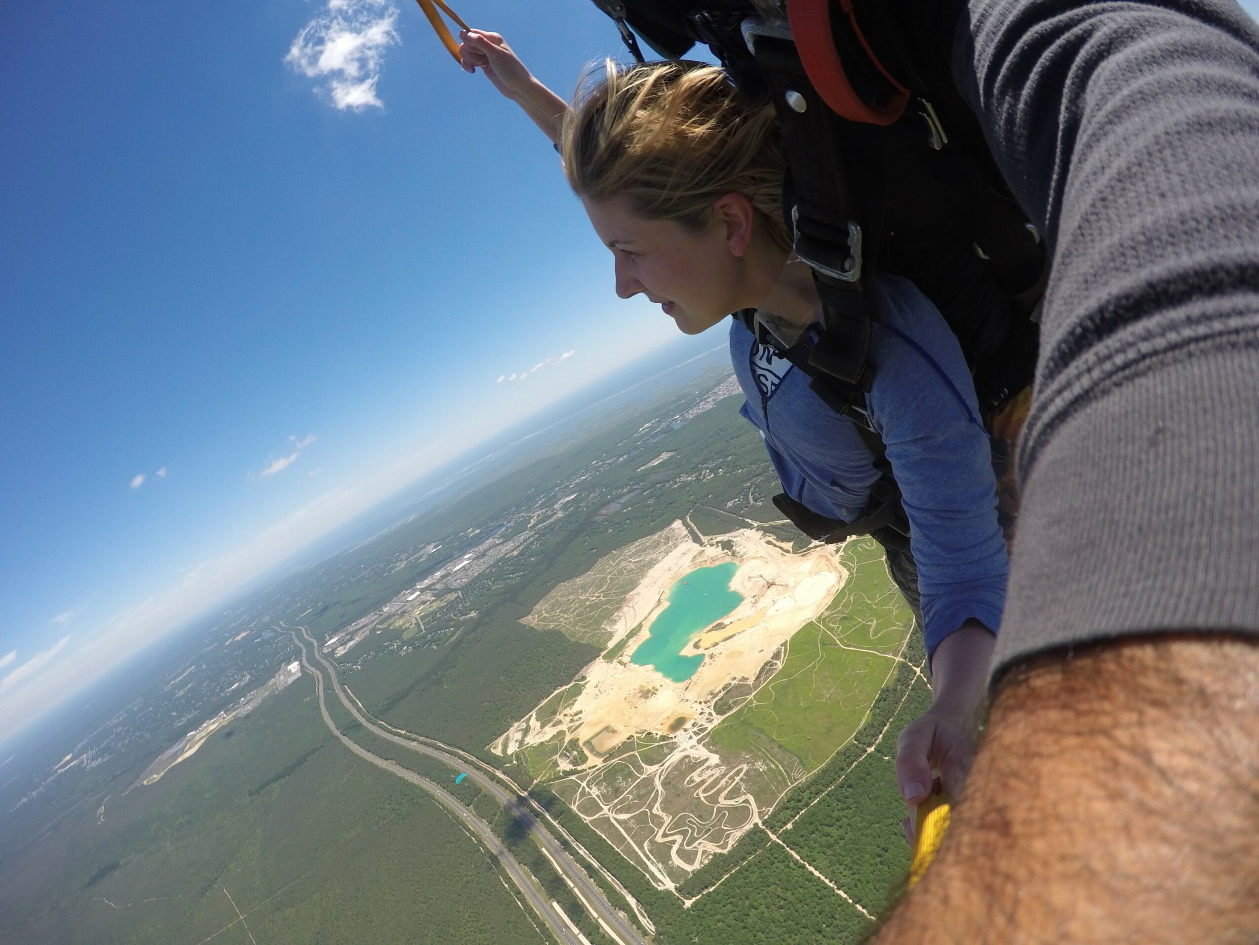 Person skydiving with another person, view of landscape with a lake and roads below, blue sky, clouds.