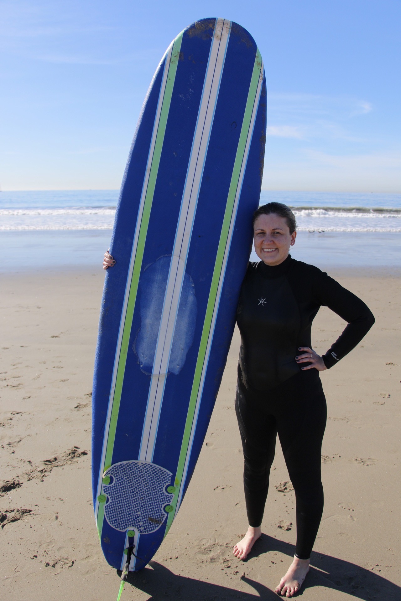Person in wetsuit standing on beach next to a large surfboard, holding it upright, with ocean and sky in background.