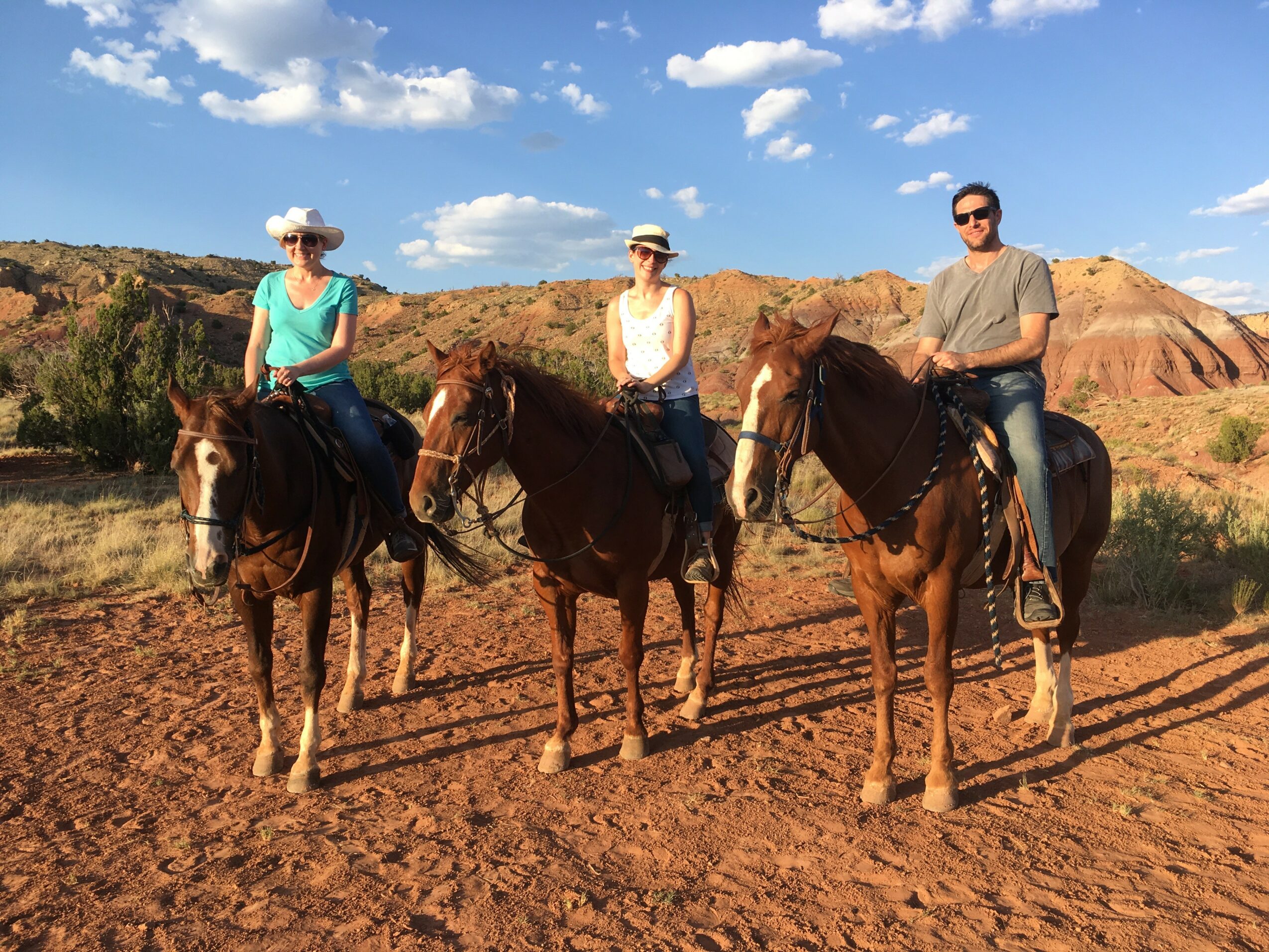 Three people on horseback in a desert landscape with red soil and hills, under a blue sky with clouds.