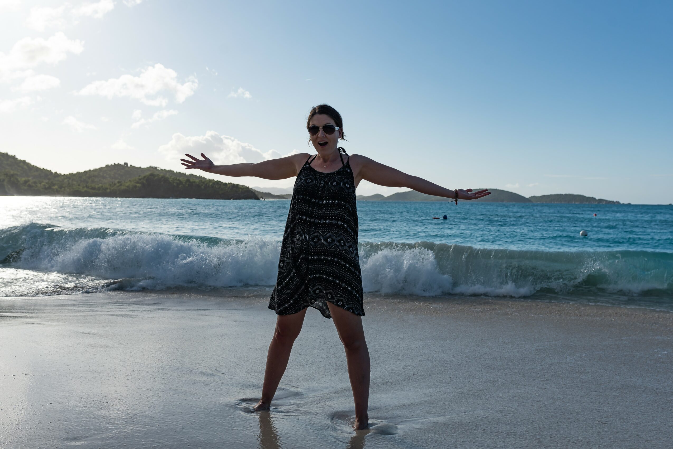 Woman standing on beach with arms outstretched, ocean waves behind her, wearing sunglasses and a patterned dress.