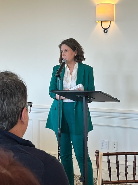 Woman standing at a podium speaking, with a microphone, in a room with a wall-mounted light fixture, audience members visible.