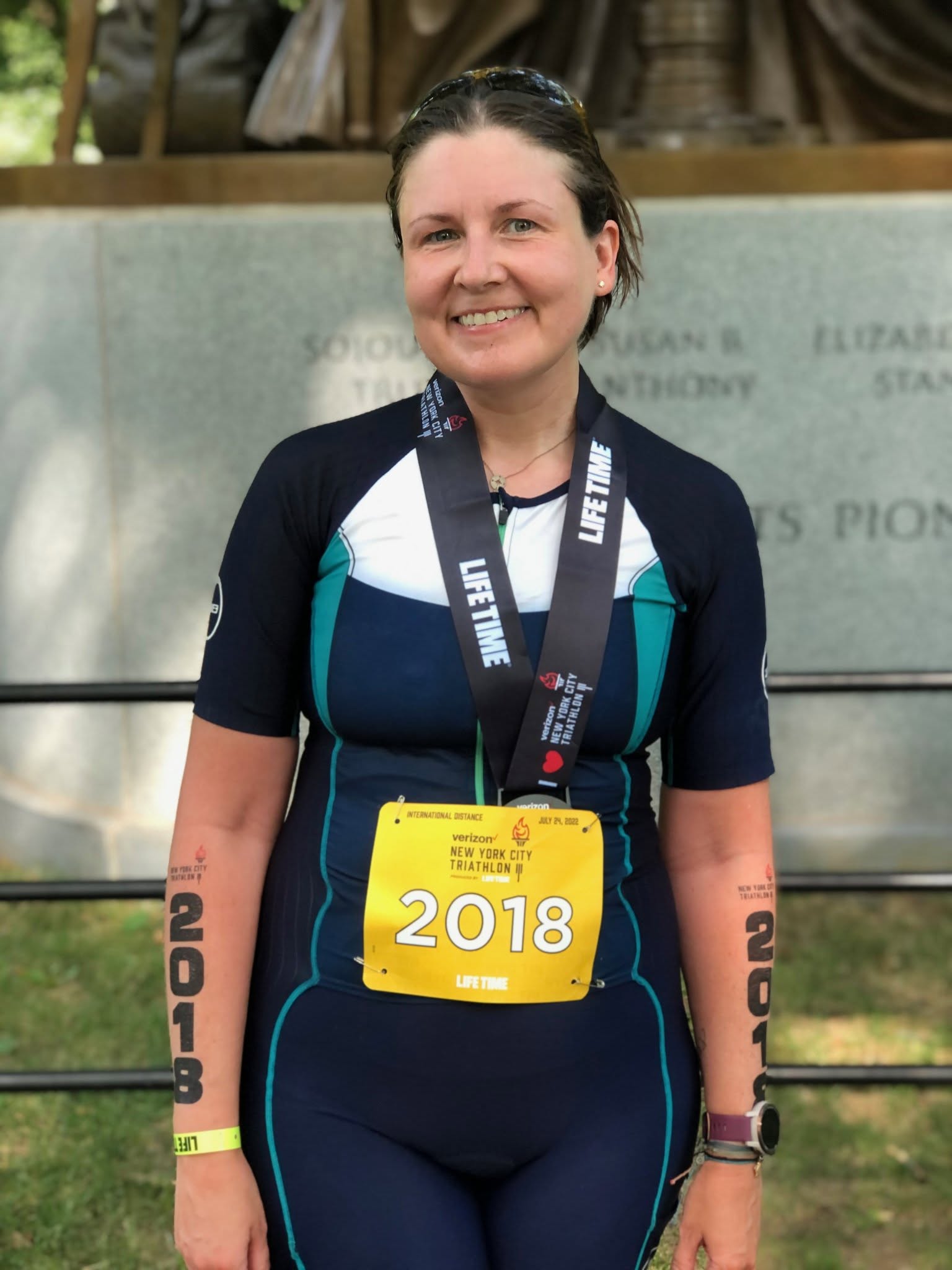 Smiling woman in athletic gear with a yellow race bib, standing outdoors, wearing a medal and a watch.