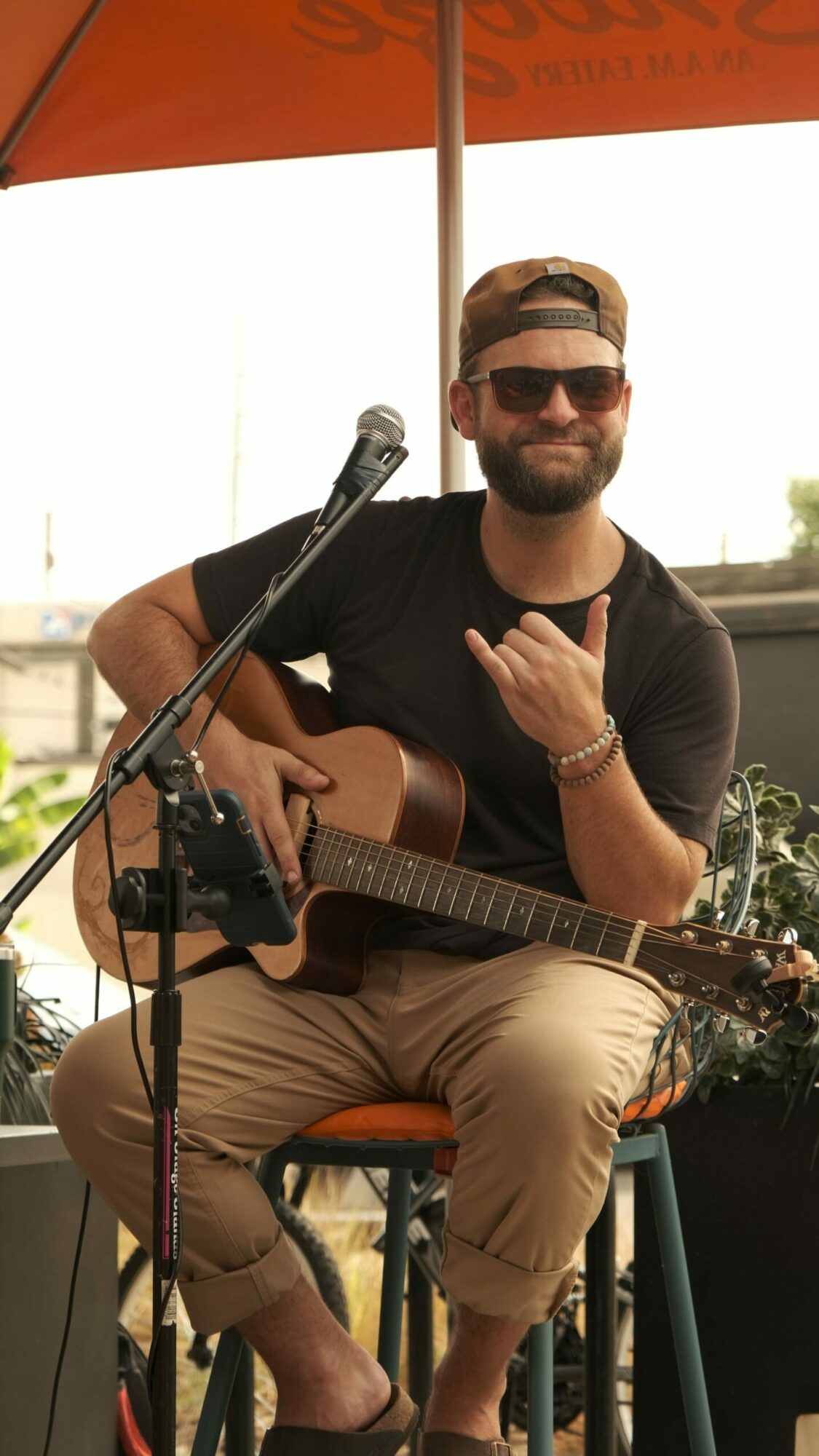 Man wearing sunglasses playing guitar at an outdoor event, smiling, with a microphone and orange banner nearby.