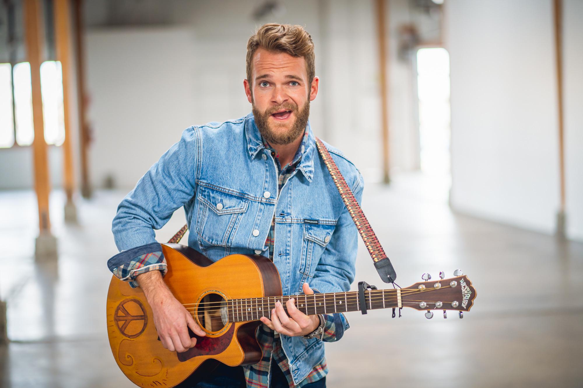 Man with a beard and denim jacket playing an acoustic guitar indoors, smiling, with a bright background.
