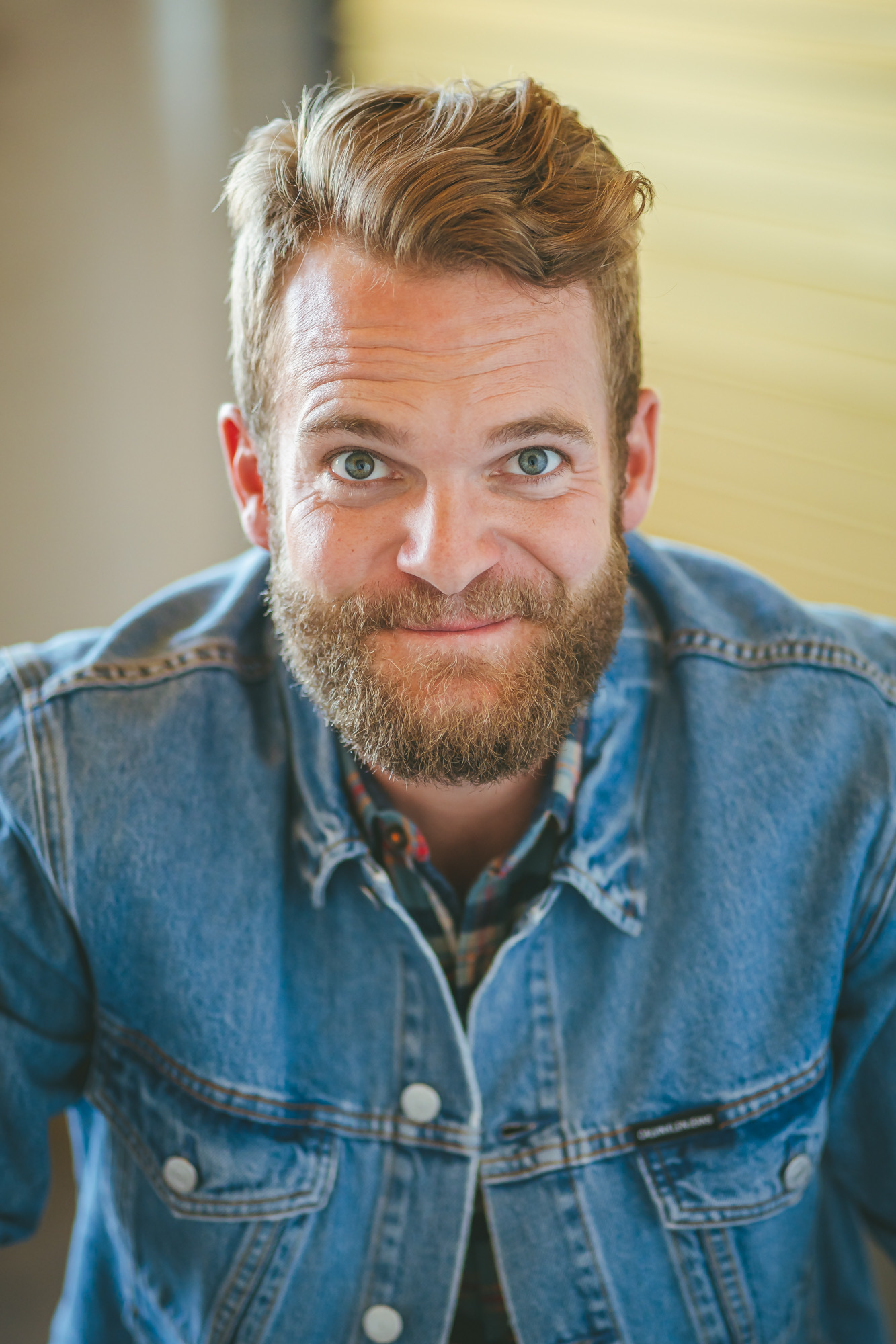 Man with a beard and short, wavy hair smiling, wearing a denim jacket, indoors with blurred background.