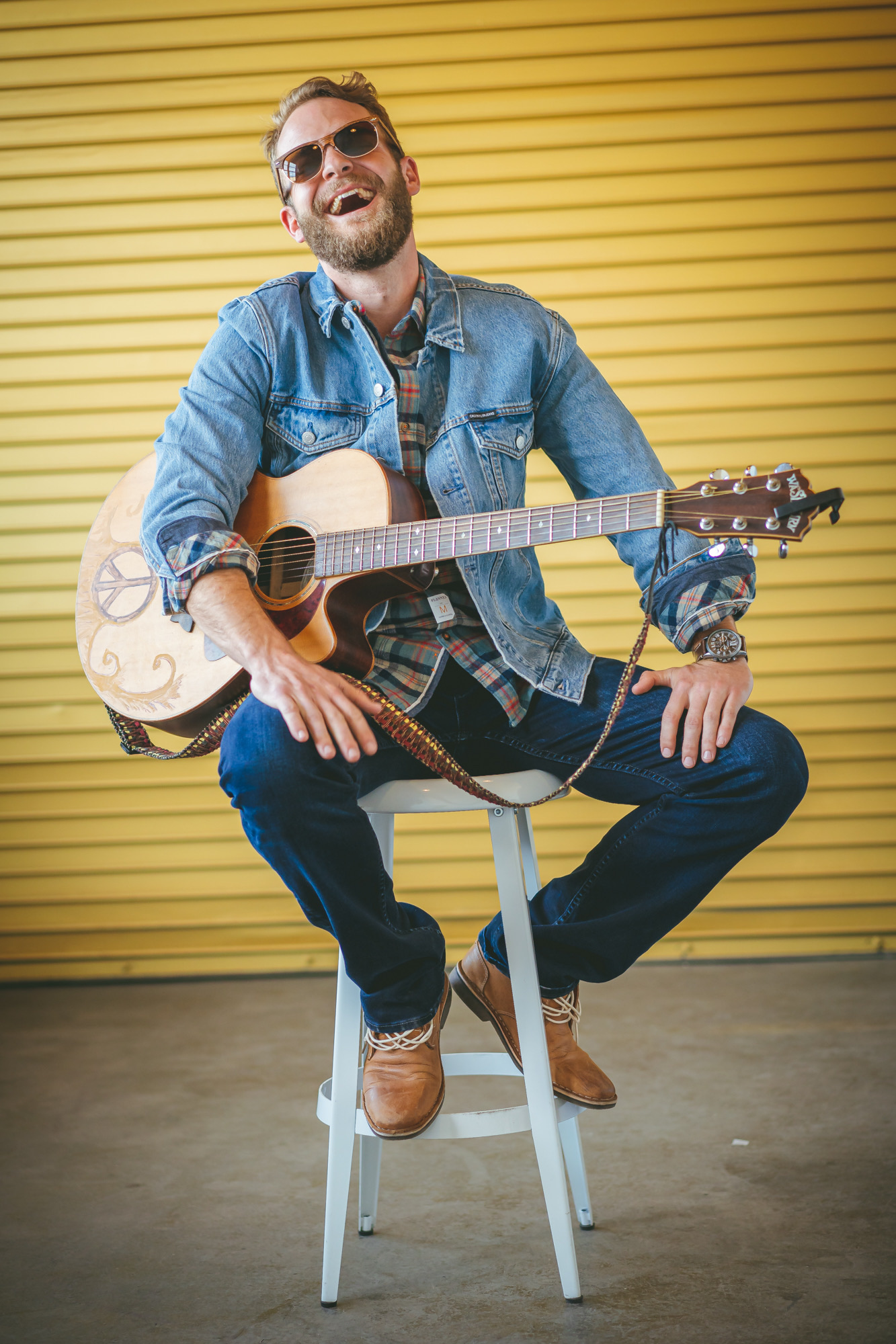 Man with sunglasses, beard, and denim jacket playing an acoustic guitar, sitting on a stool against a yellow background.