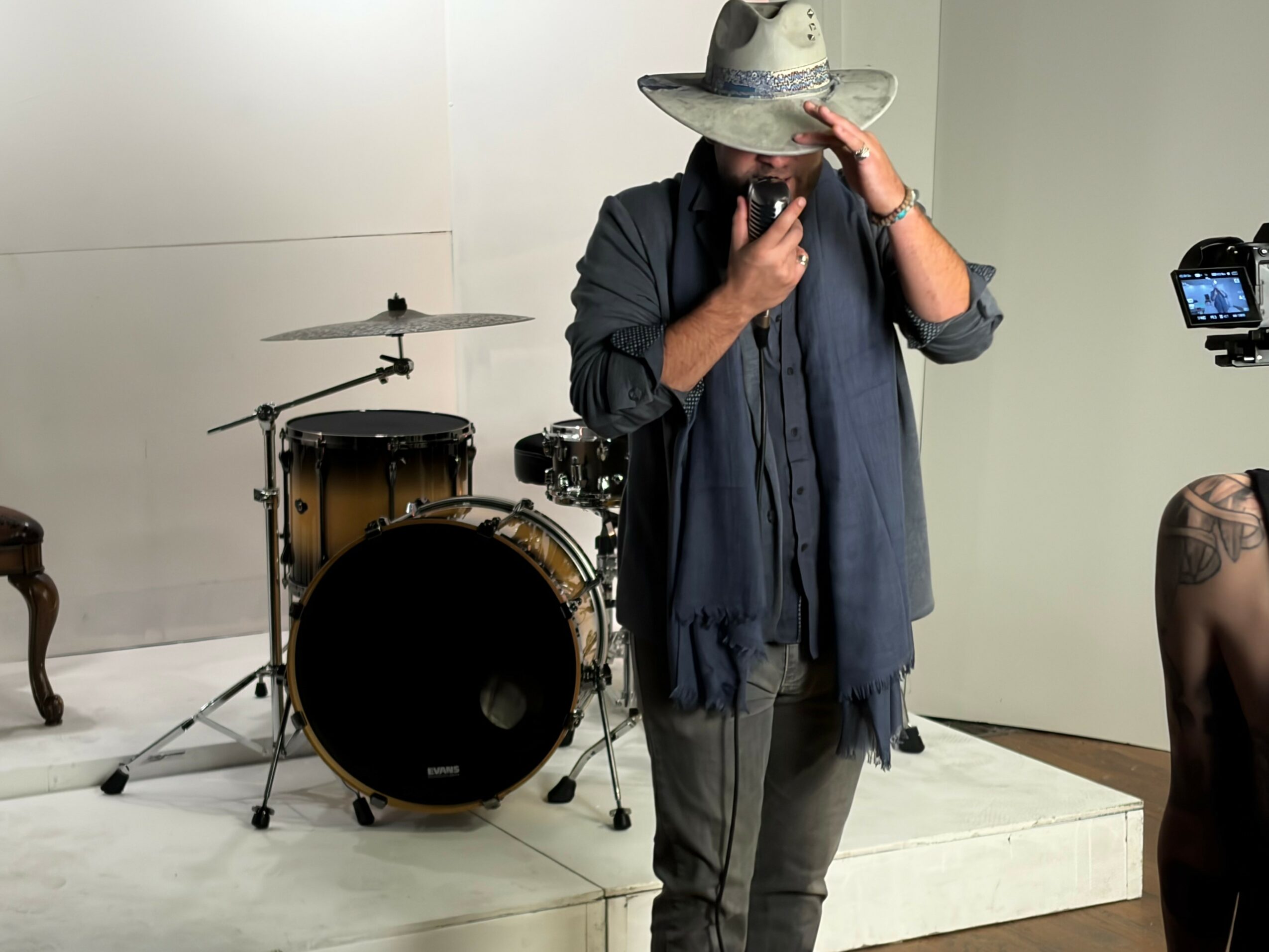Person wearing a large hat, dark jacket, and scarf, standing near musical instruments, with a camera on a tripod nearby.