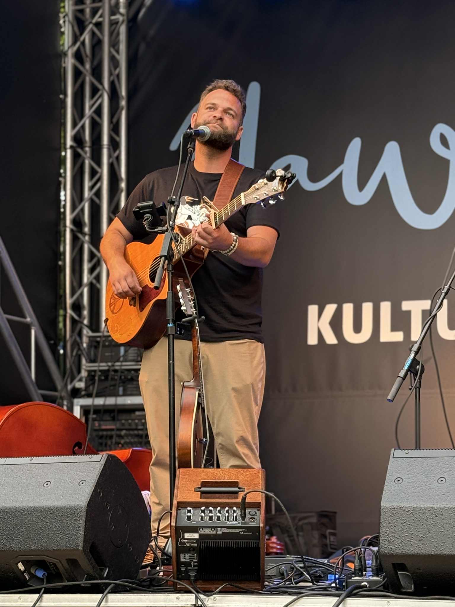 Man with a beard playing an acoustic guitar on stage, singing into a microphone, with stage equipment and a black backdrop.