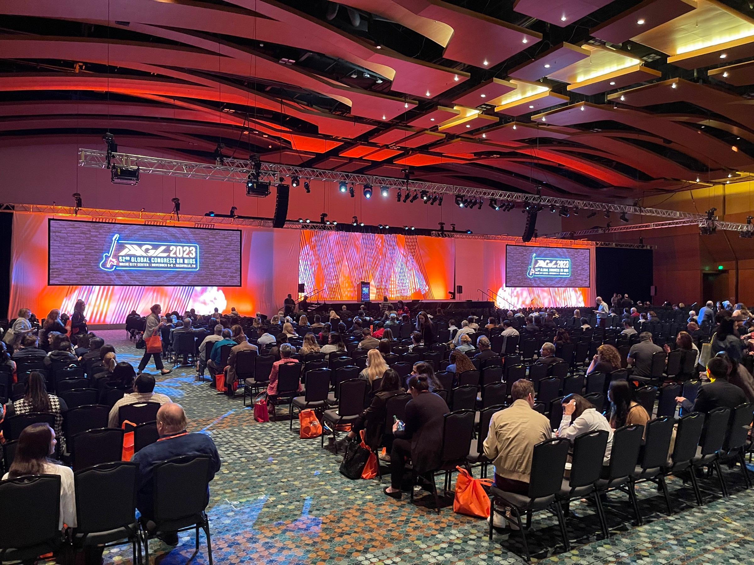 Large conference room filled with seated audience facing a stage with two large screens, colorful lighting, and a decorated ceiling.