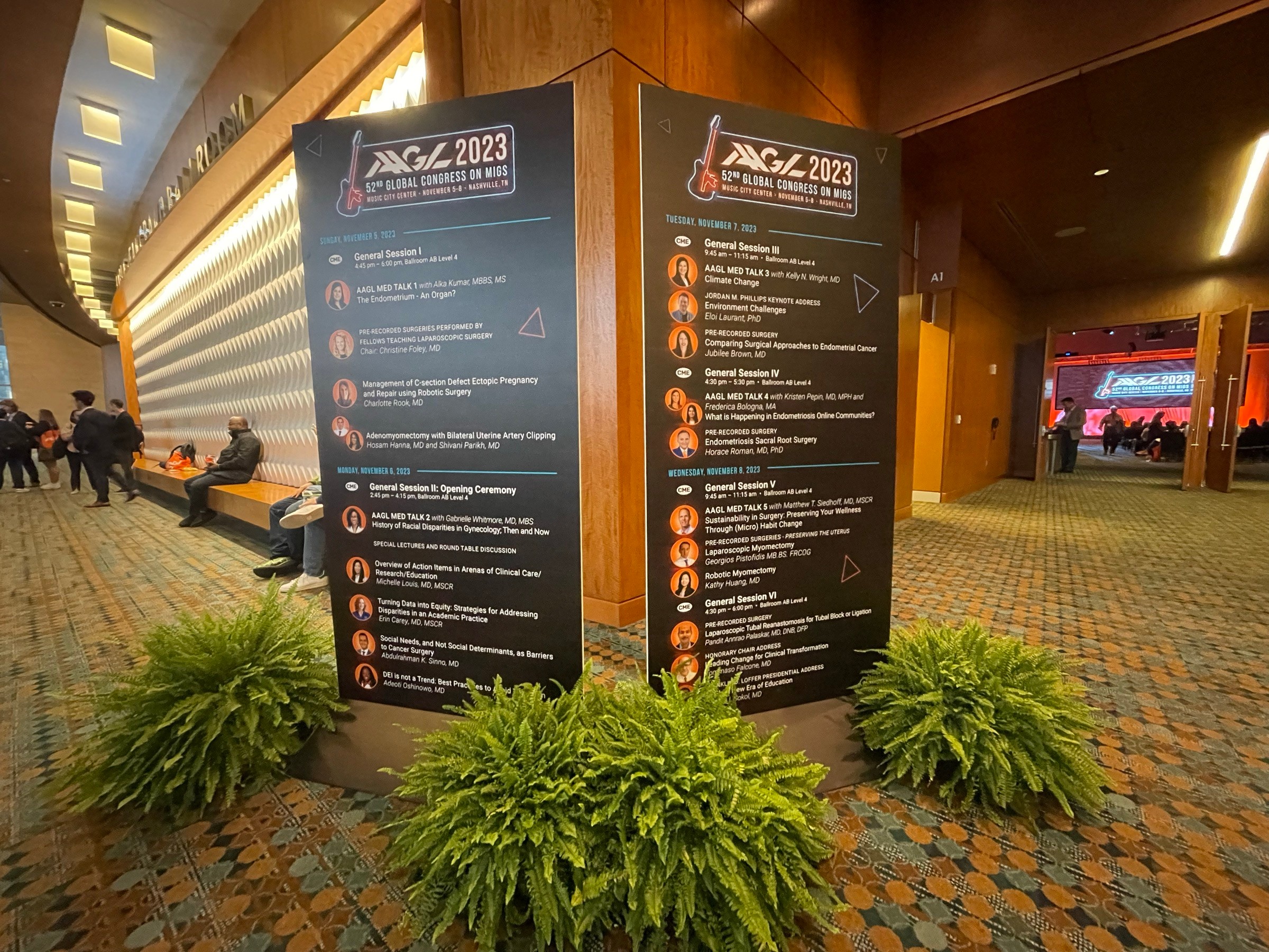 Two large vertical signs with event schedules, surrounded by green plants, in a spacious indoor area with seating and people.