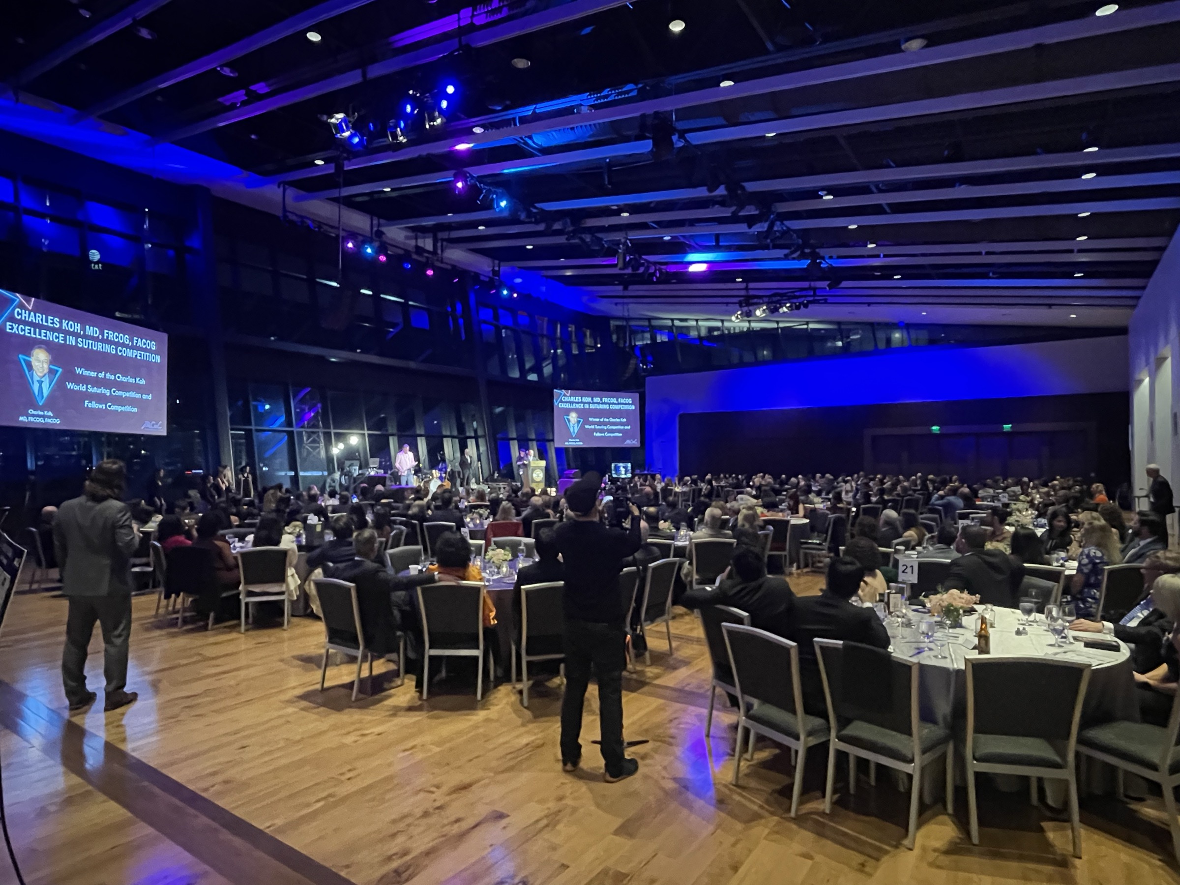 Large conference hall filled with round tables and chairs, audience seated, stage with screens and blue lighting, high ceiling.