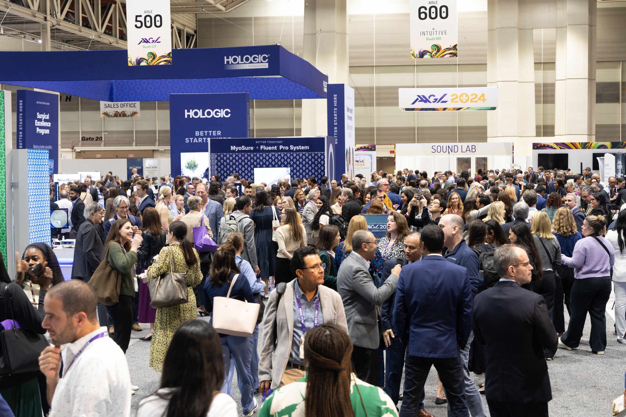 Large crowd of people at an indoor exhibition with booths and banners, some engaging in conversations and exploring displays.
