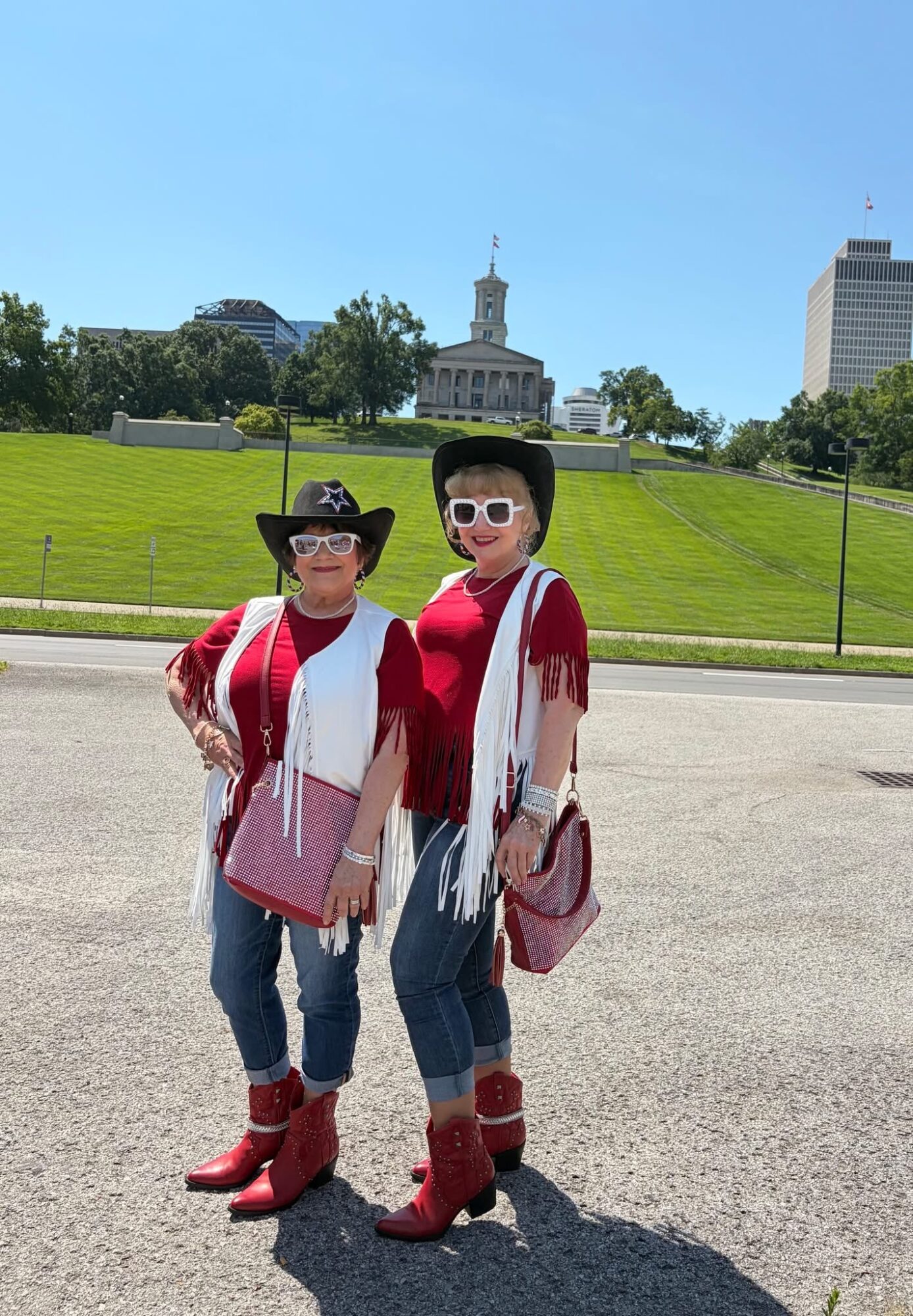 Two women stand outdoors on a sunny day, wearing cowboy hats, sunglasses, red tops, jeans, and red boots, with a green hill and buildings in the background.