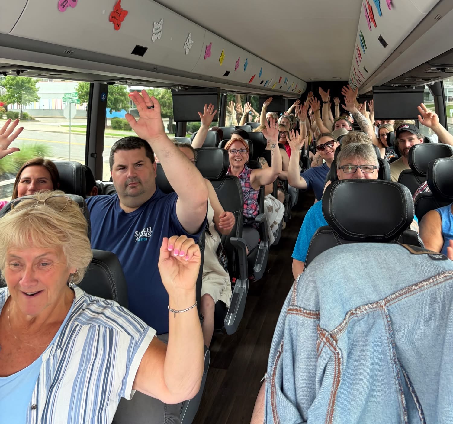 Group of people on a bus raising hands, smiling, with windows showing trees outside, and colorful decorations overhead.