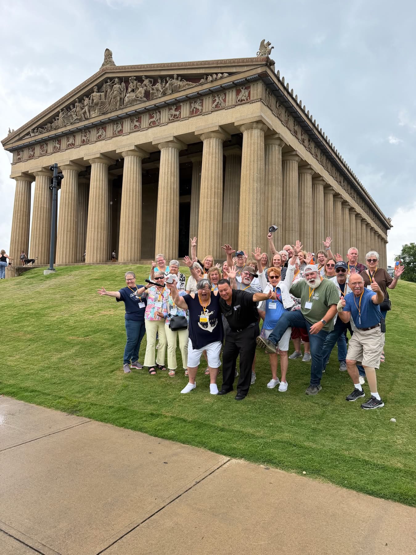 Group of people in front of a classical Greek temple with columns and detailed pediment, on a grassy area.