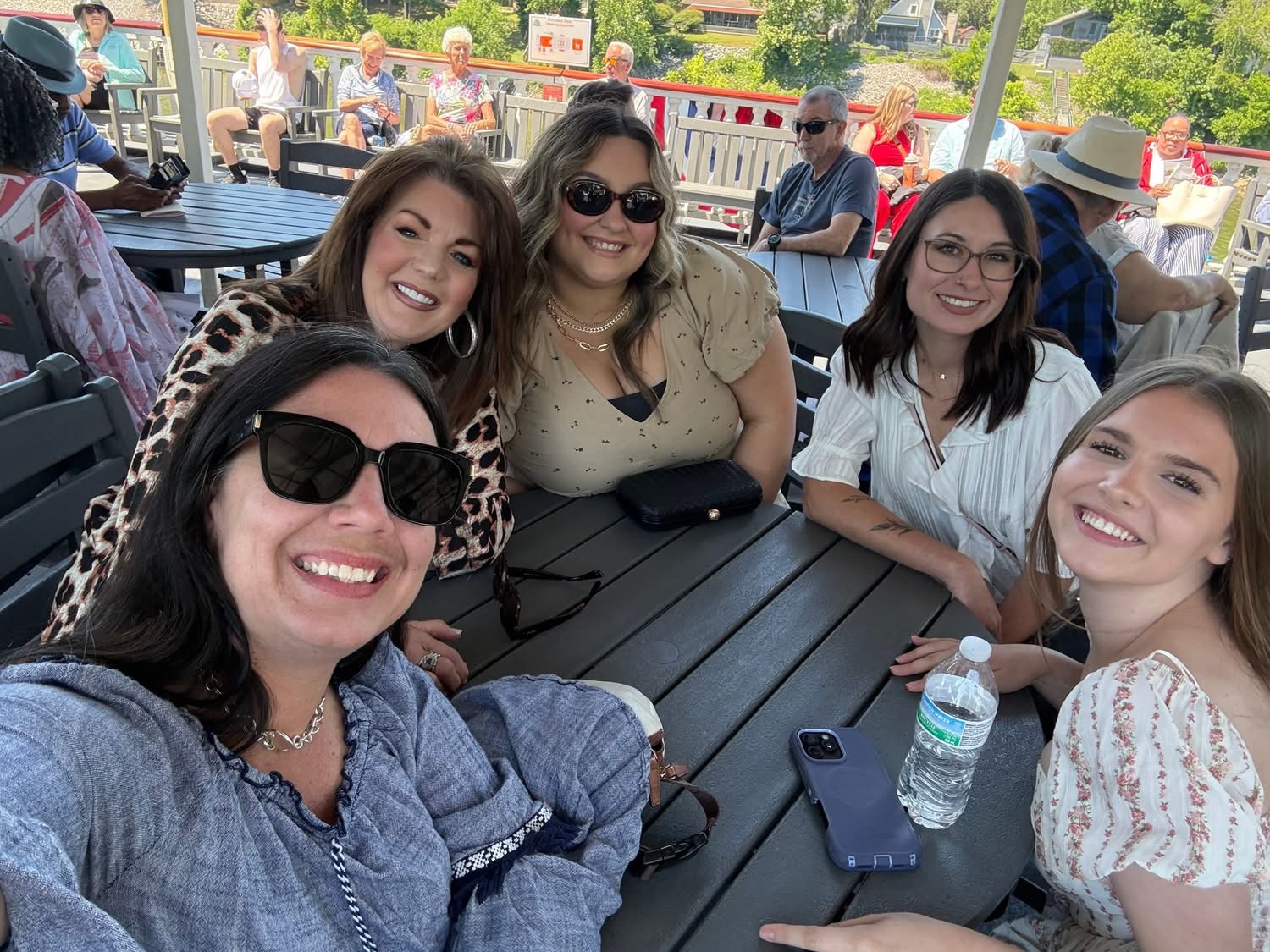 Five women sitting at a table outdoors, smiling, with drinks and sunglasses, enjoying a sunny day.