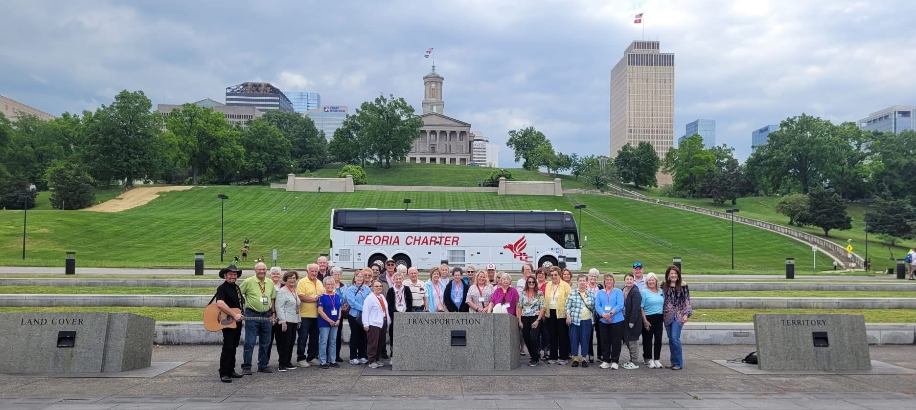 Group of people standing in front of a bus with a city park and buildings in the background.