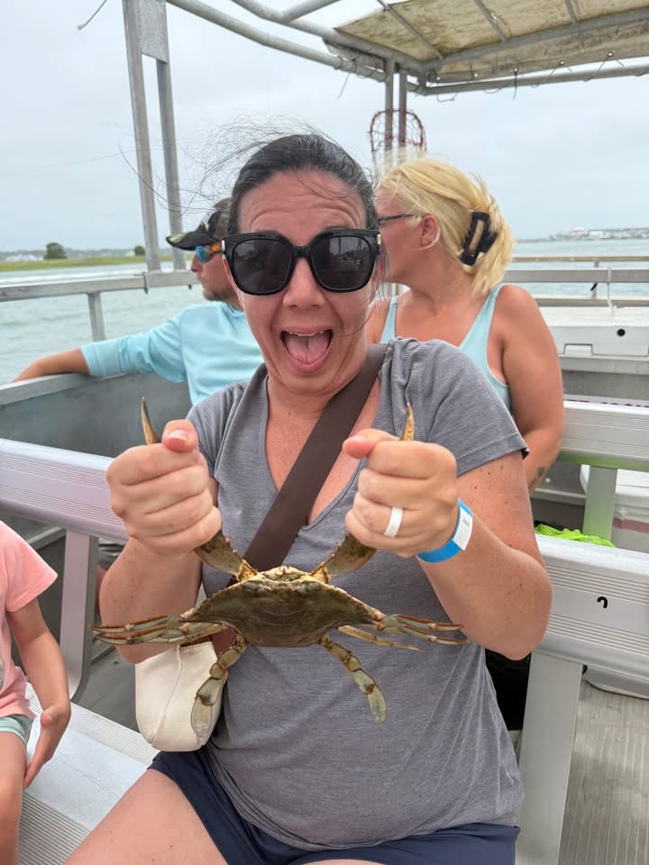 Woman with sunglasses smiling and pointing, holding a crab, outdoors on a boat or dock.