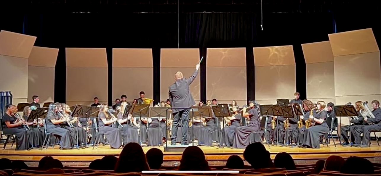 Orchestra performing on stage with conductor, audience visible in foreground, stage with large beige panels in background.