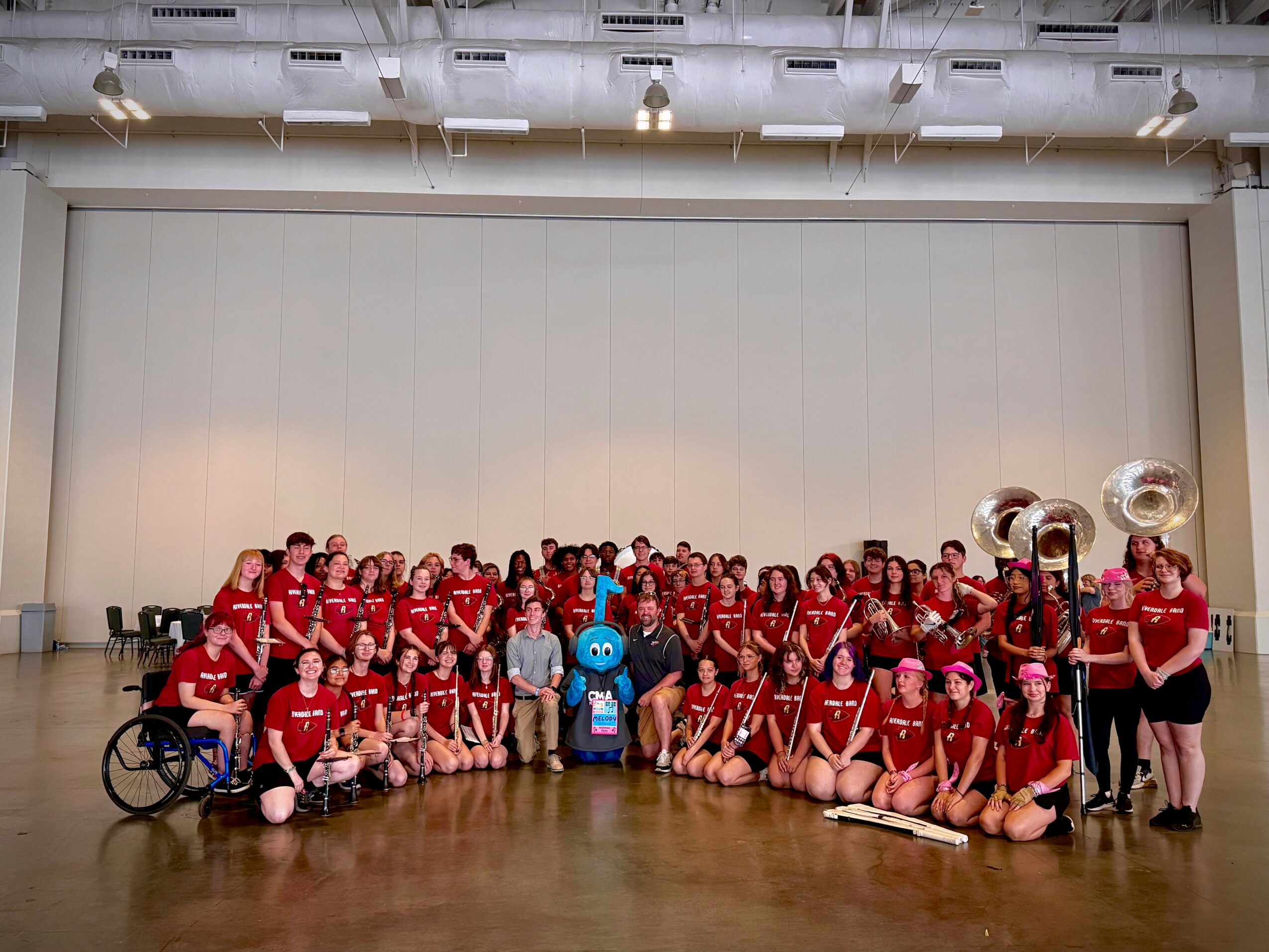 Group of children and adults in red shirts, some holding musical instruments, on stage with a blue mascot, in a large hall.