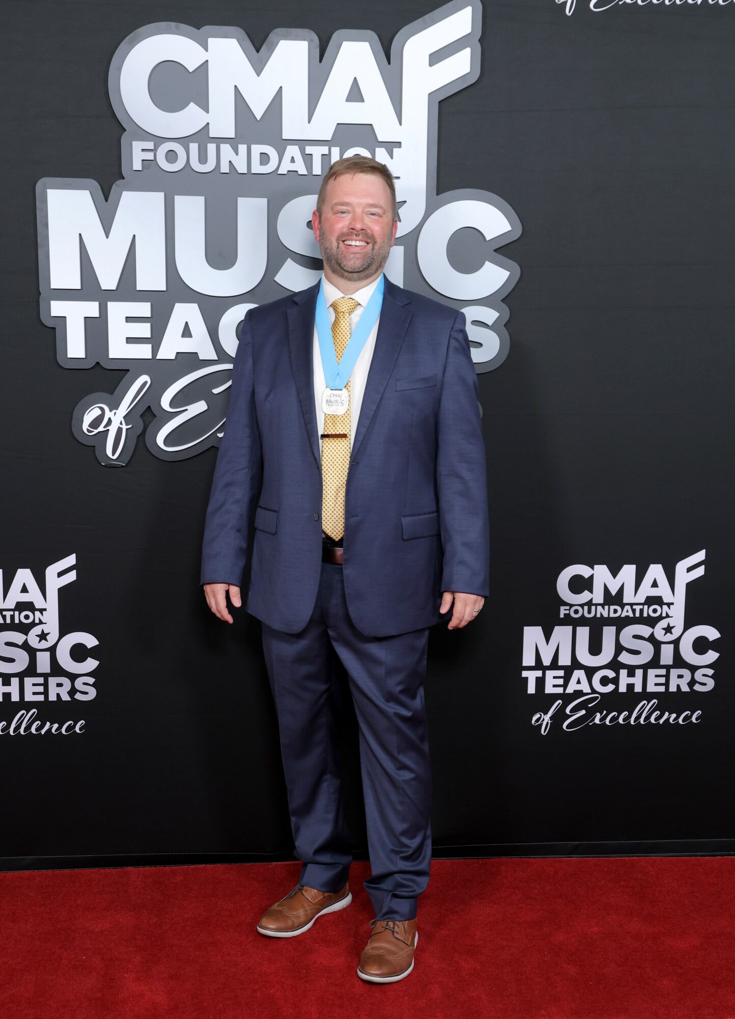 Man in a suit standing on red carpet at CMAF event, smiling, background with event logo, dark backdrop, and text.