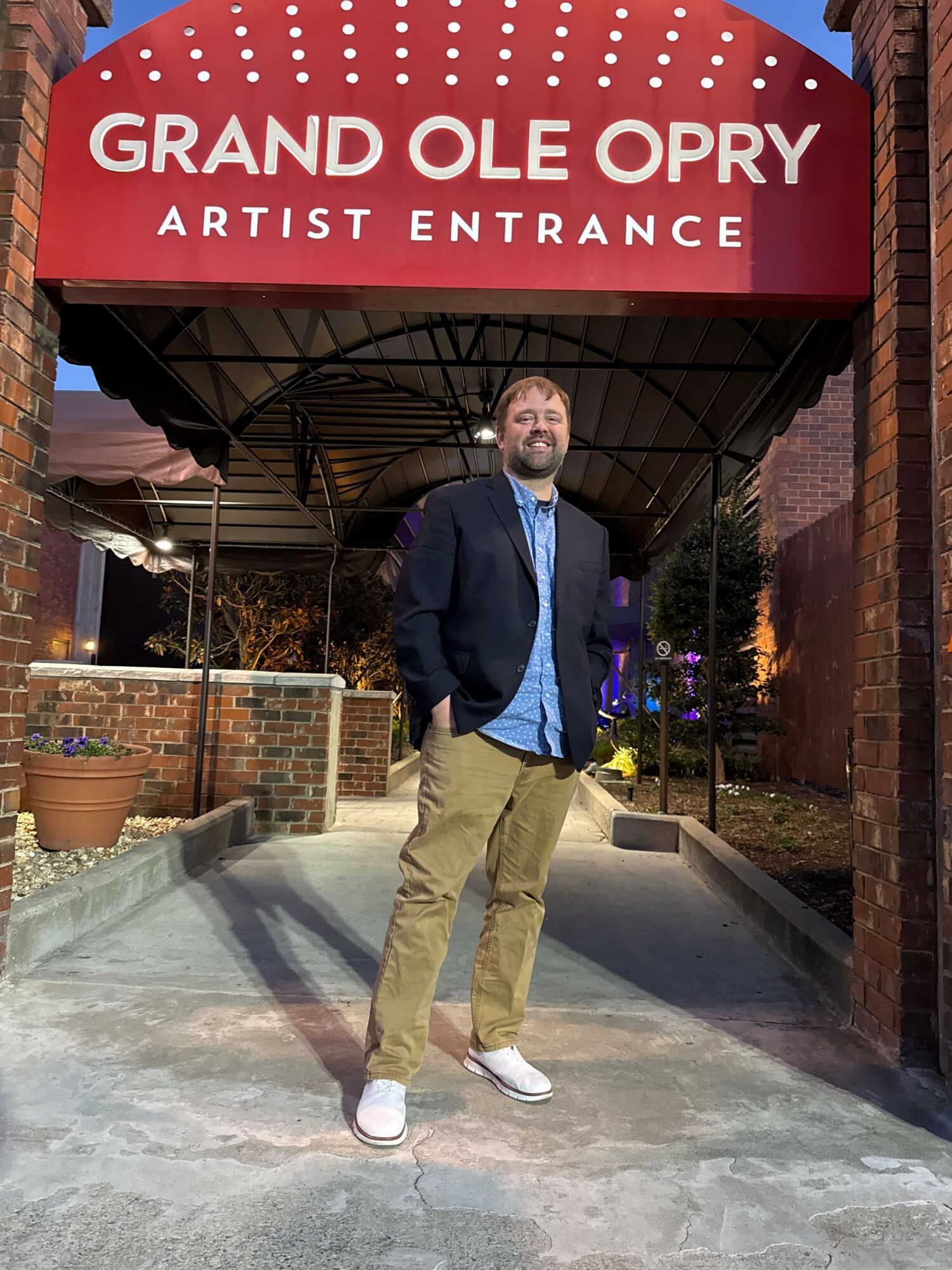 Man standing outside under a sign that reads 'Grand Ole Opry Artist Entrance' at night.