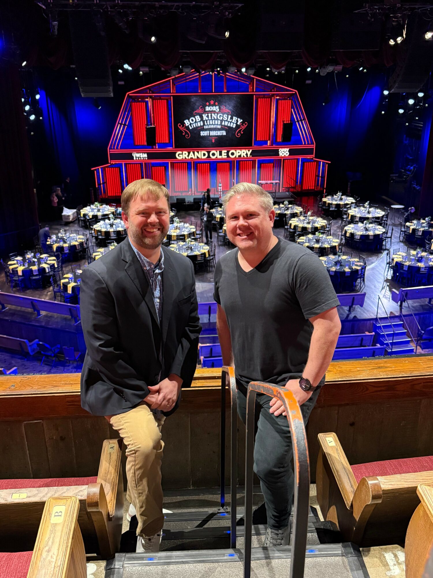 Two men stand on a balcony overlooking a stage with a large screen and tables, smiling at the camera.