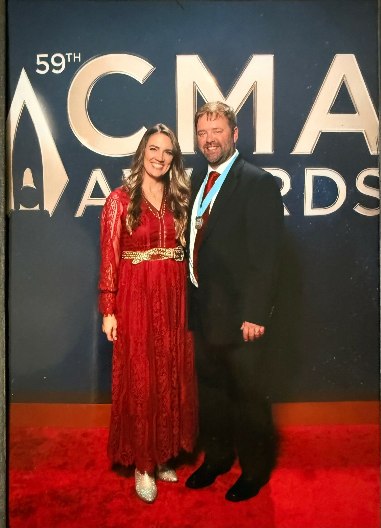Two people standing in front of a large sign reading '59th CMA Awards,' smiling, with a dark background and red carpet.