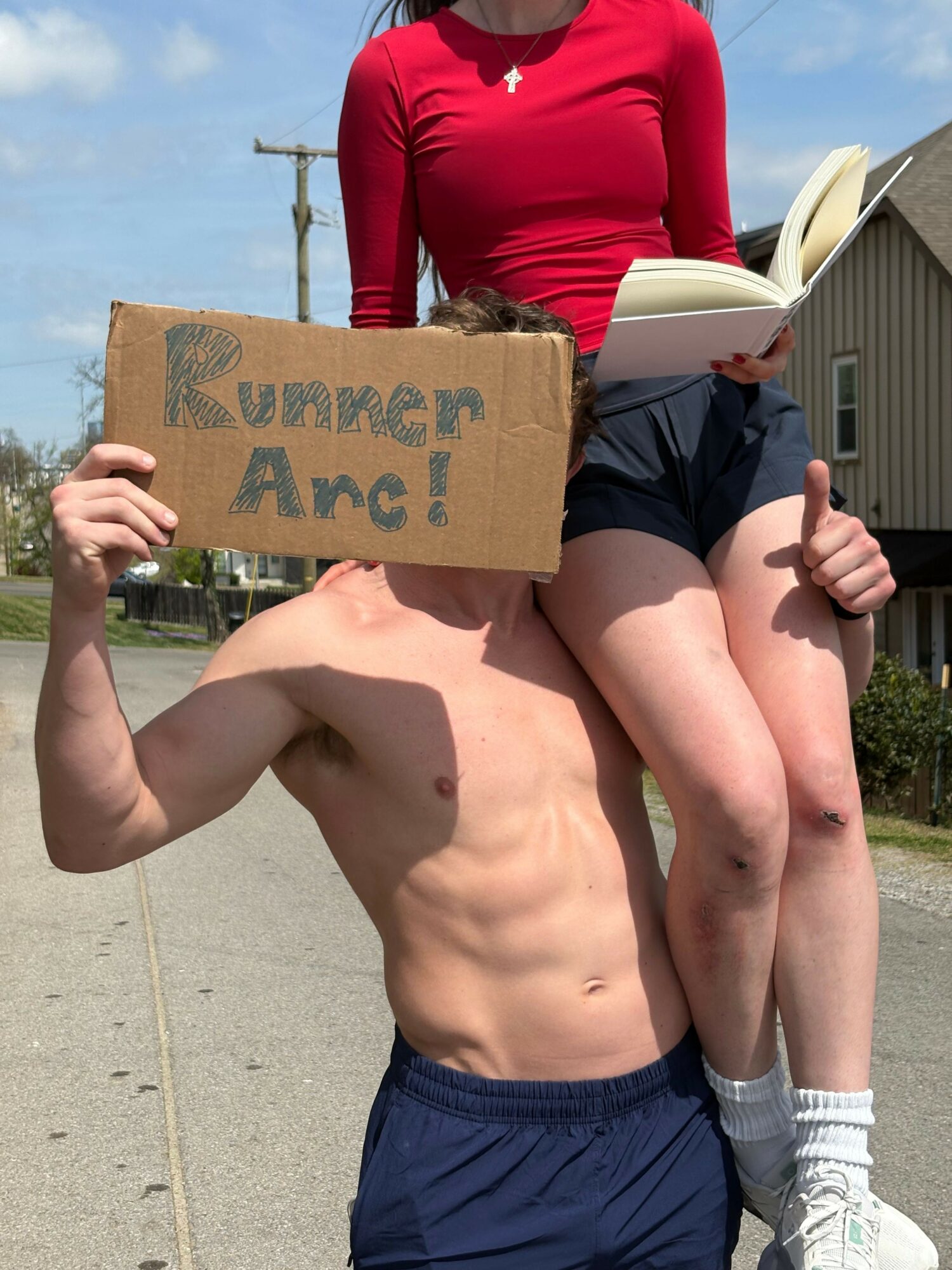 Person holding a cardboard sign with handwritten text, standing outdoors with another person sitting nearby, both in casual clothing.