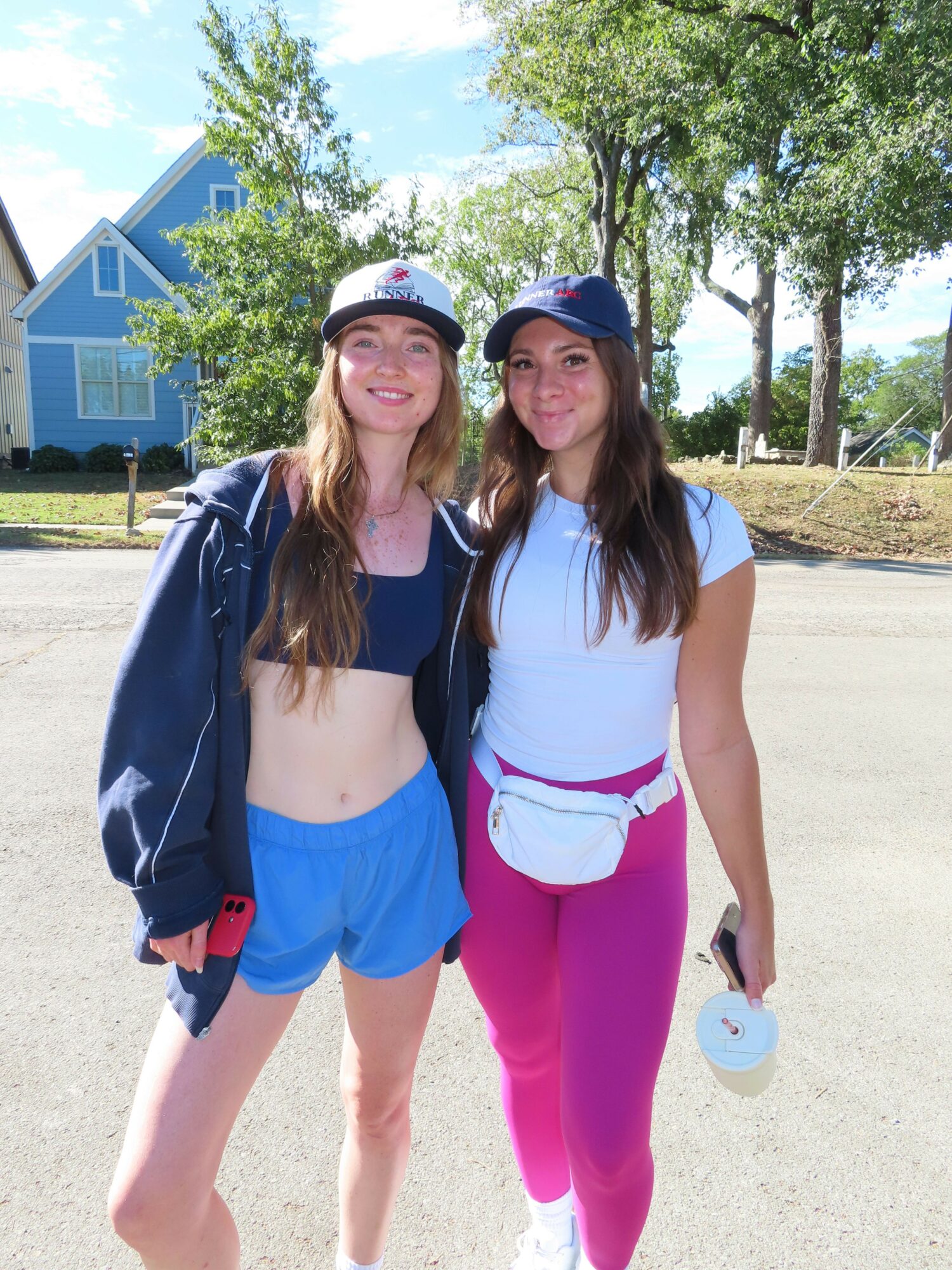 Two young women standing outdoors on a sunny day, one in pink pants and white top, the other in blue shorts and a cap, smiling.