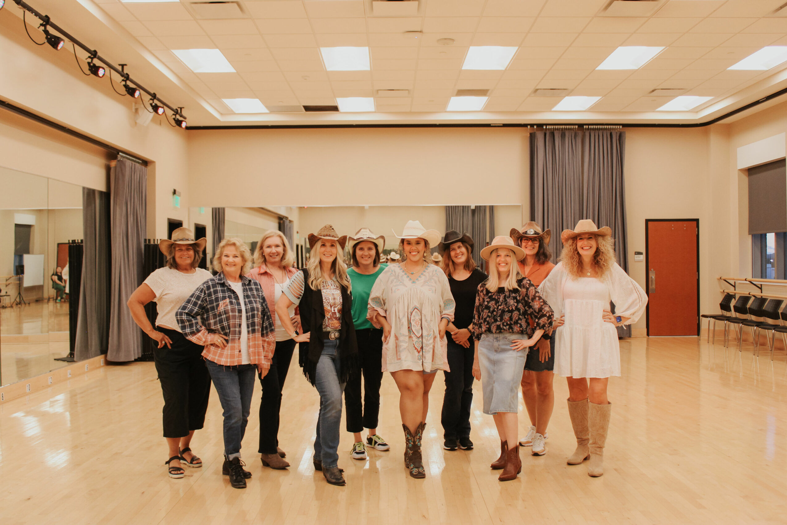 Group of nine women standing in a dance studio with wooden floor and curtains, smiling and posing for the photo.