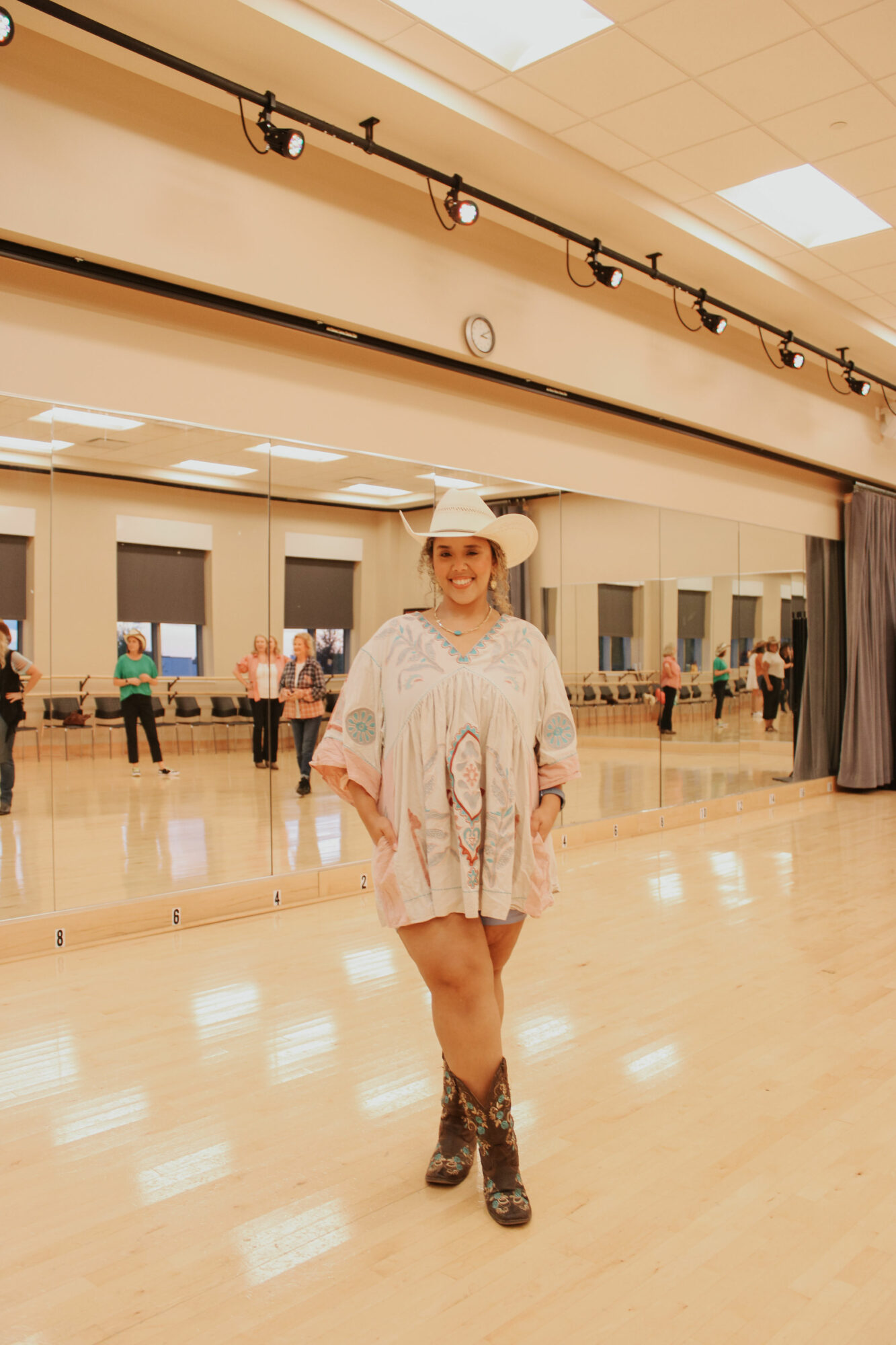 Person in a white outfit and hat standing in a dance studio with mirrors and people in the background.