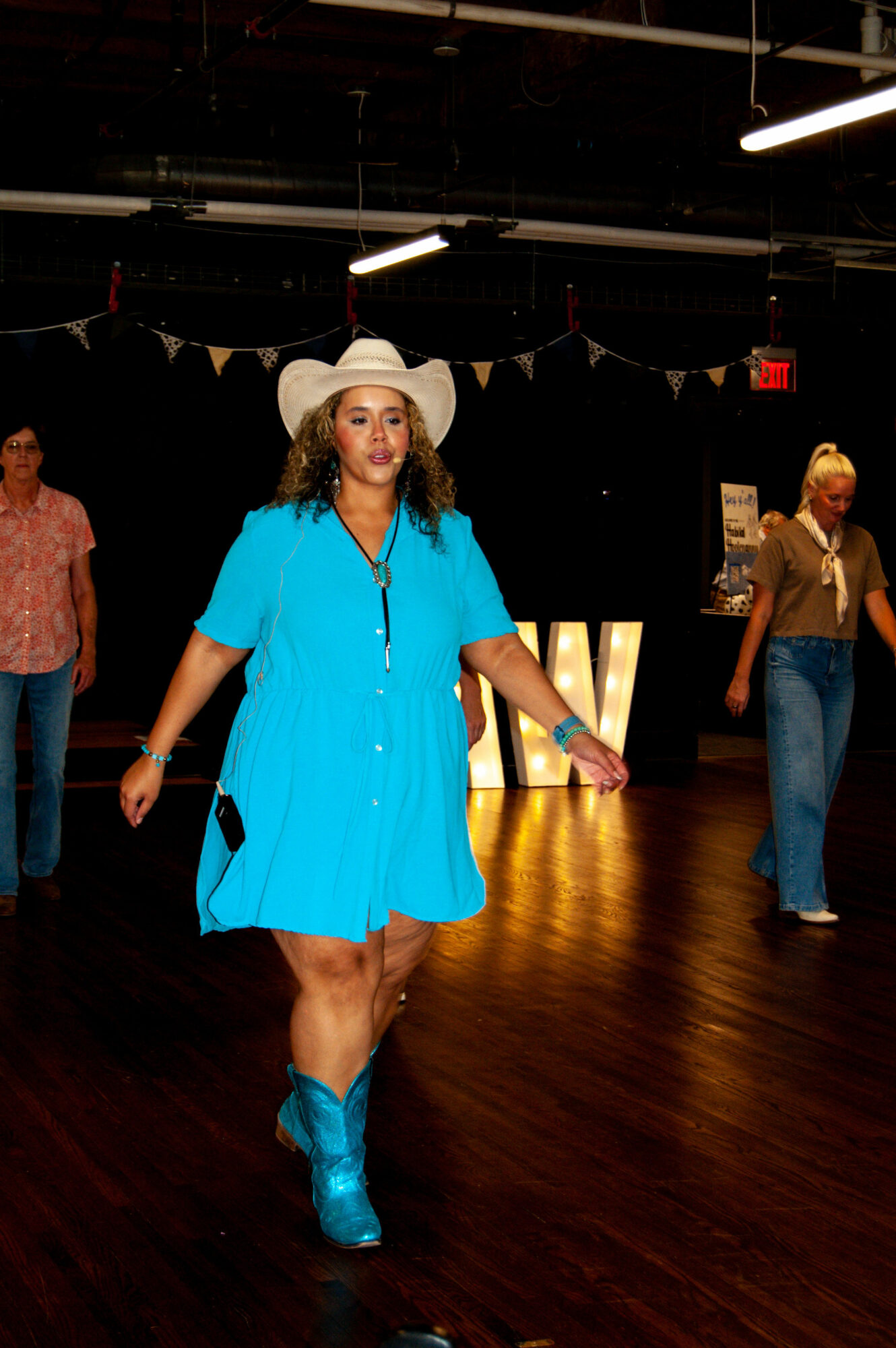 Woman in a bright blue dress and cowboy hat walking on a wooden floor, with people in the background.